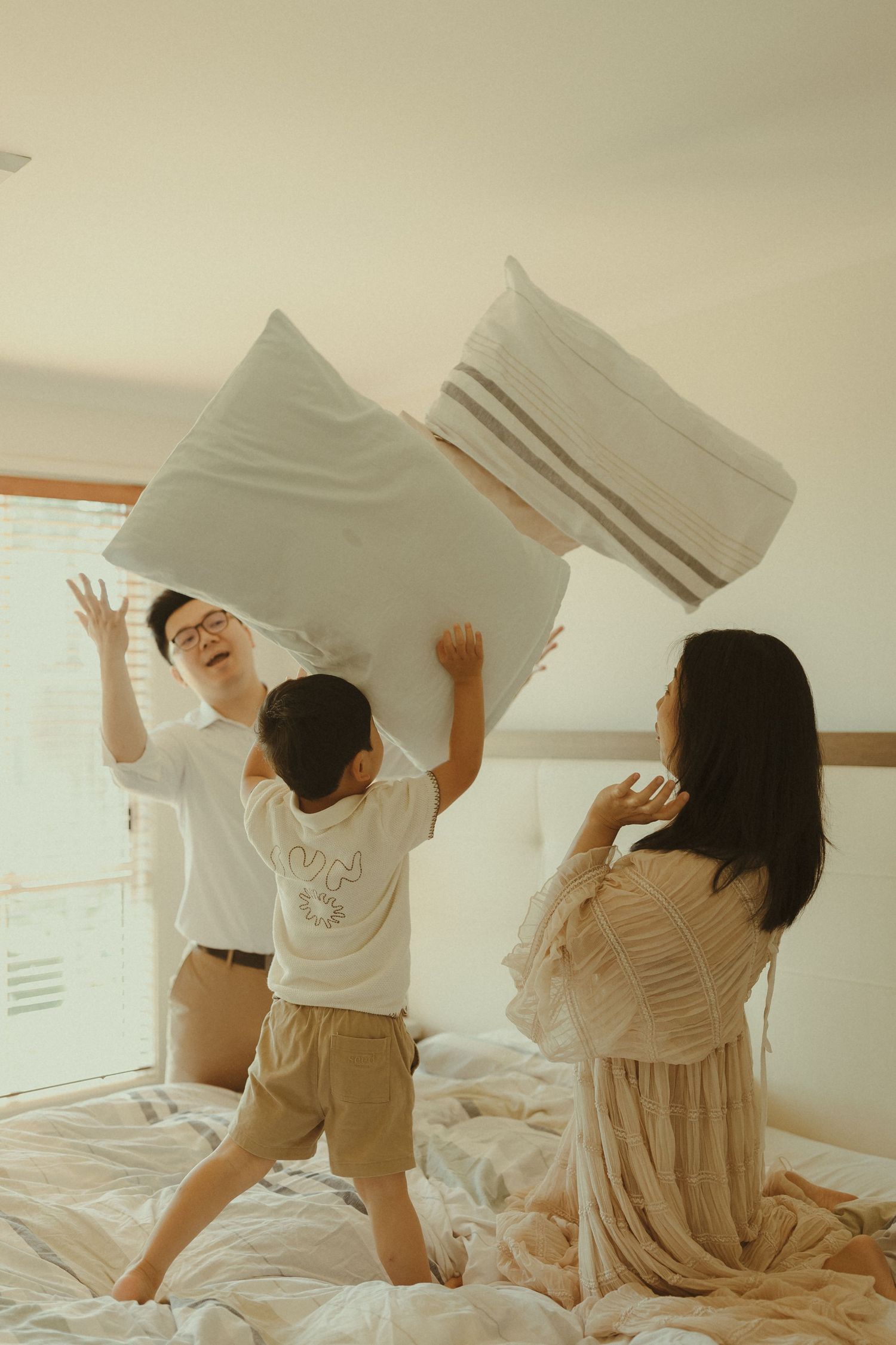 People having a pillow fight on a white bed with natural lighting.