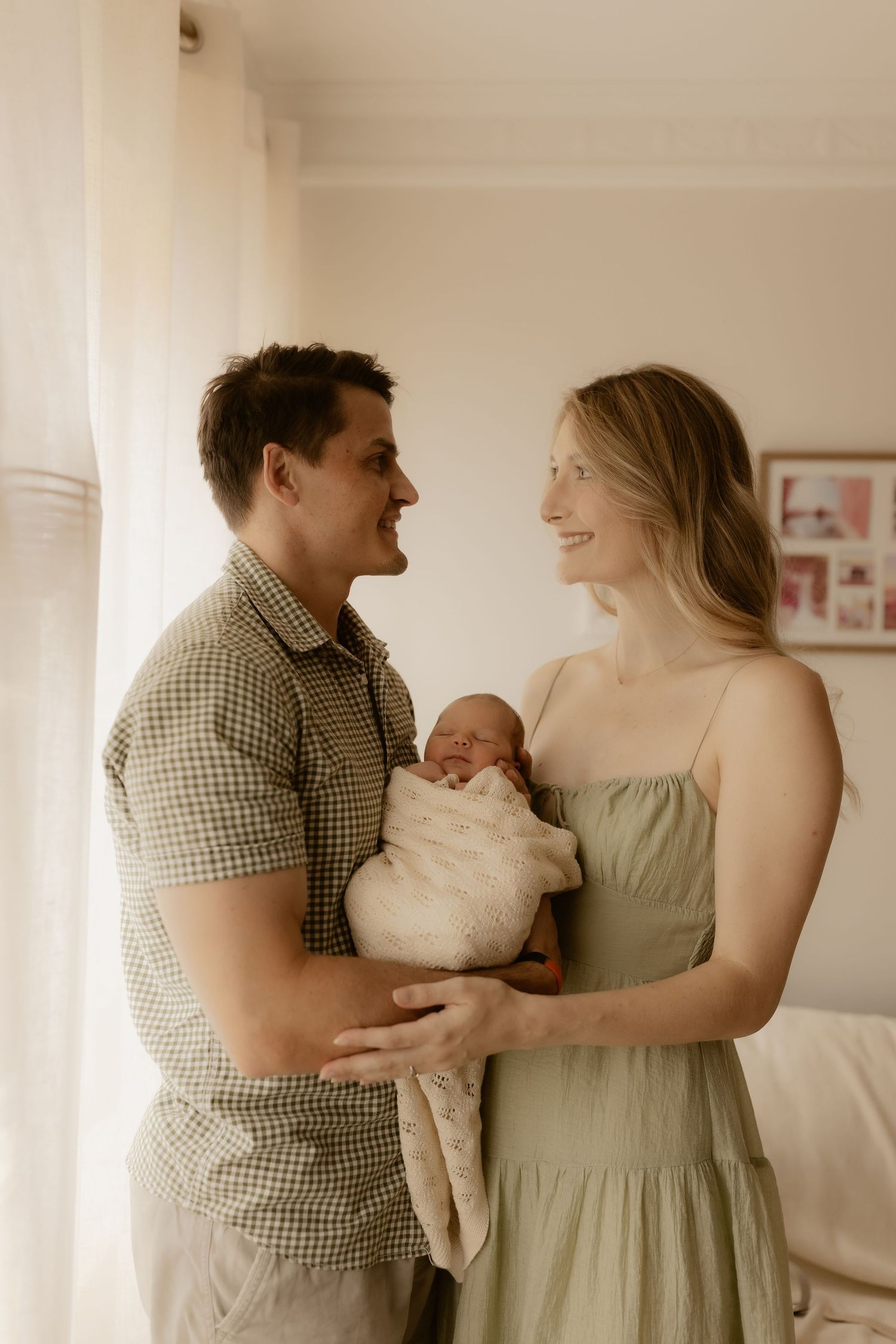 New parents sharing tender moment with swaddled newborn in sunlit room.