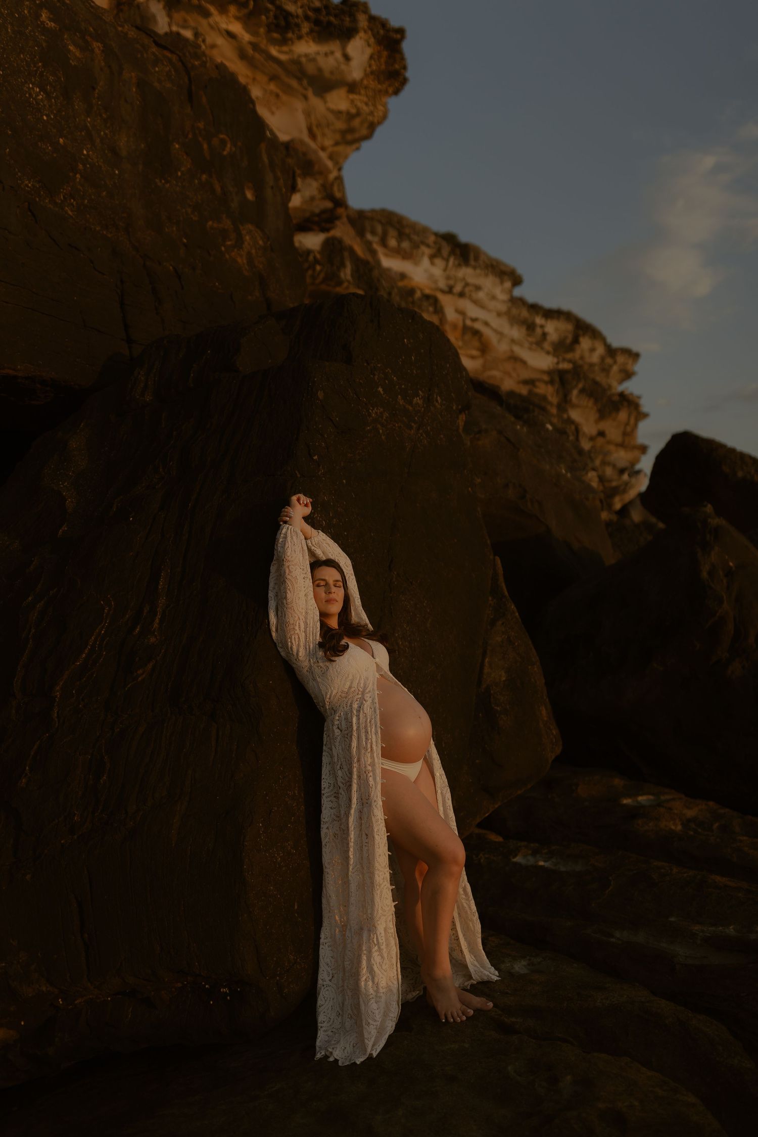 Ethereal figure in flowing white dress poses against dramatic rocky cliffs at sunset creating a dreamy silhouette.