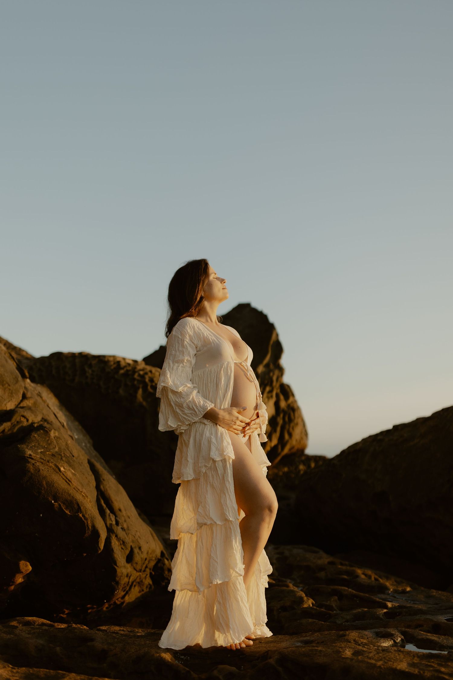 Person in ethereal white tiered dress poses on rocky cliff at sunset.