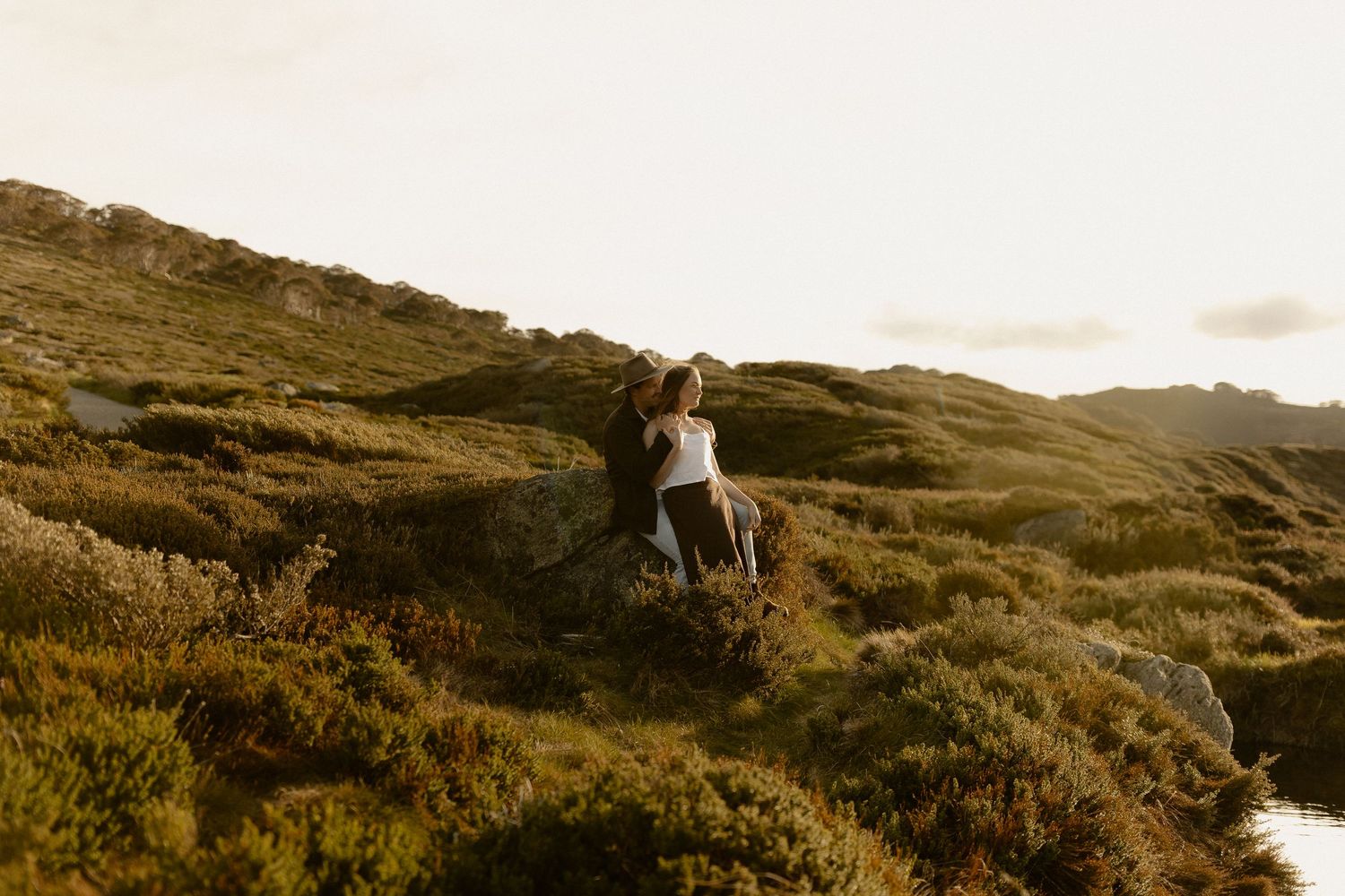 Silhouettes of two people embrace on a hilltop during sunset with mountains in the background.