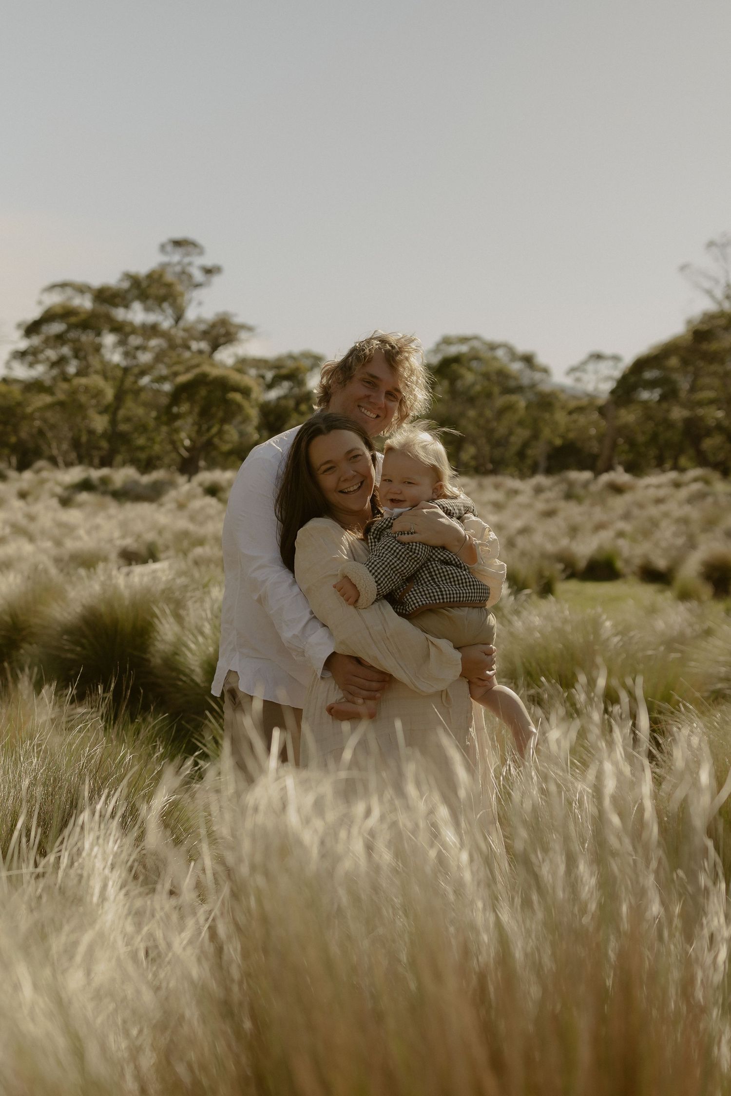 A parent and child share a tender embrace while standing in a sunlit field of tall grass.