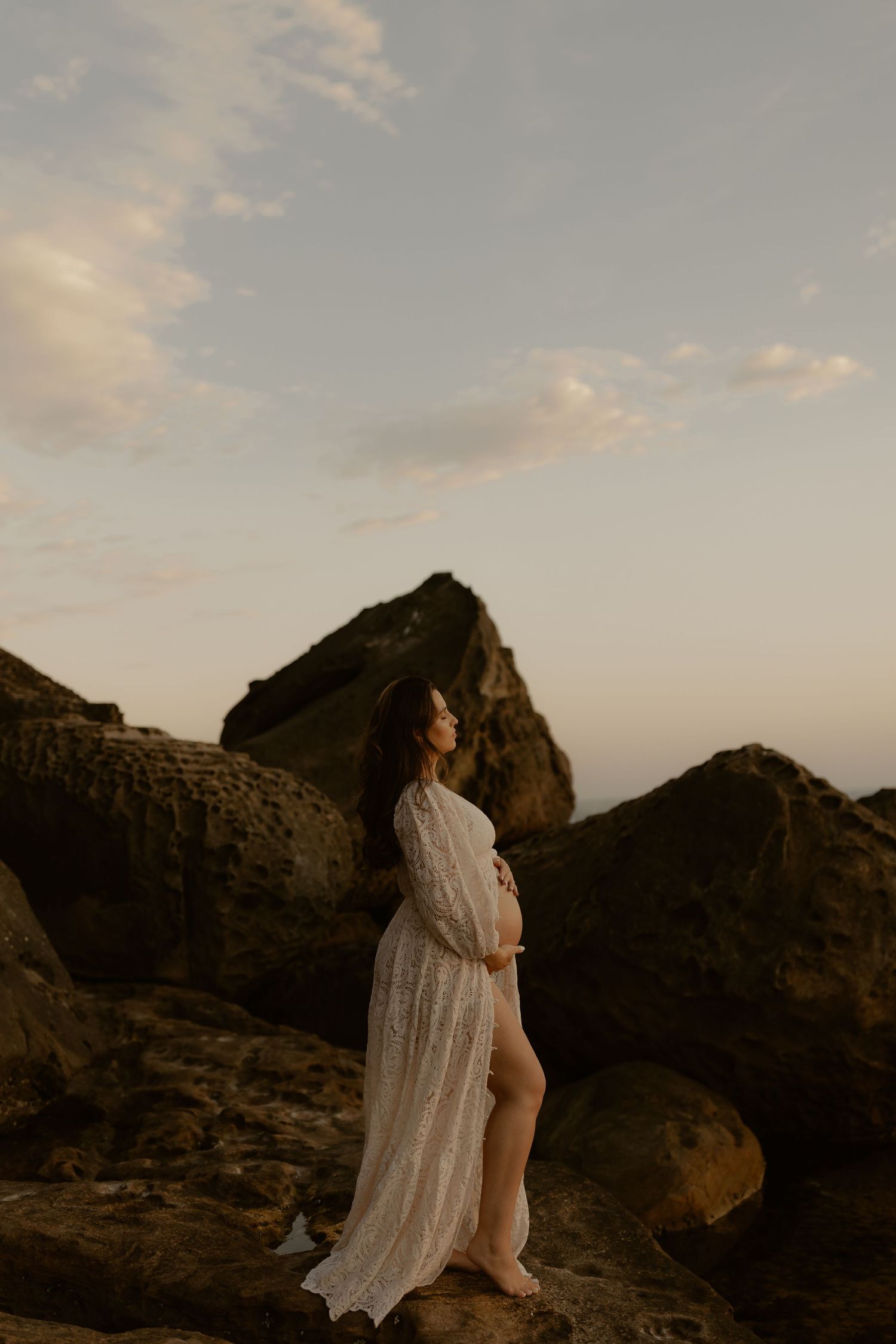 Figure in flowing white dress stands on rocky cliff at sunset with dramatic mountain silhouette in background.