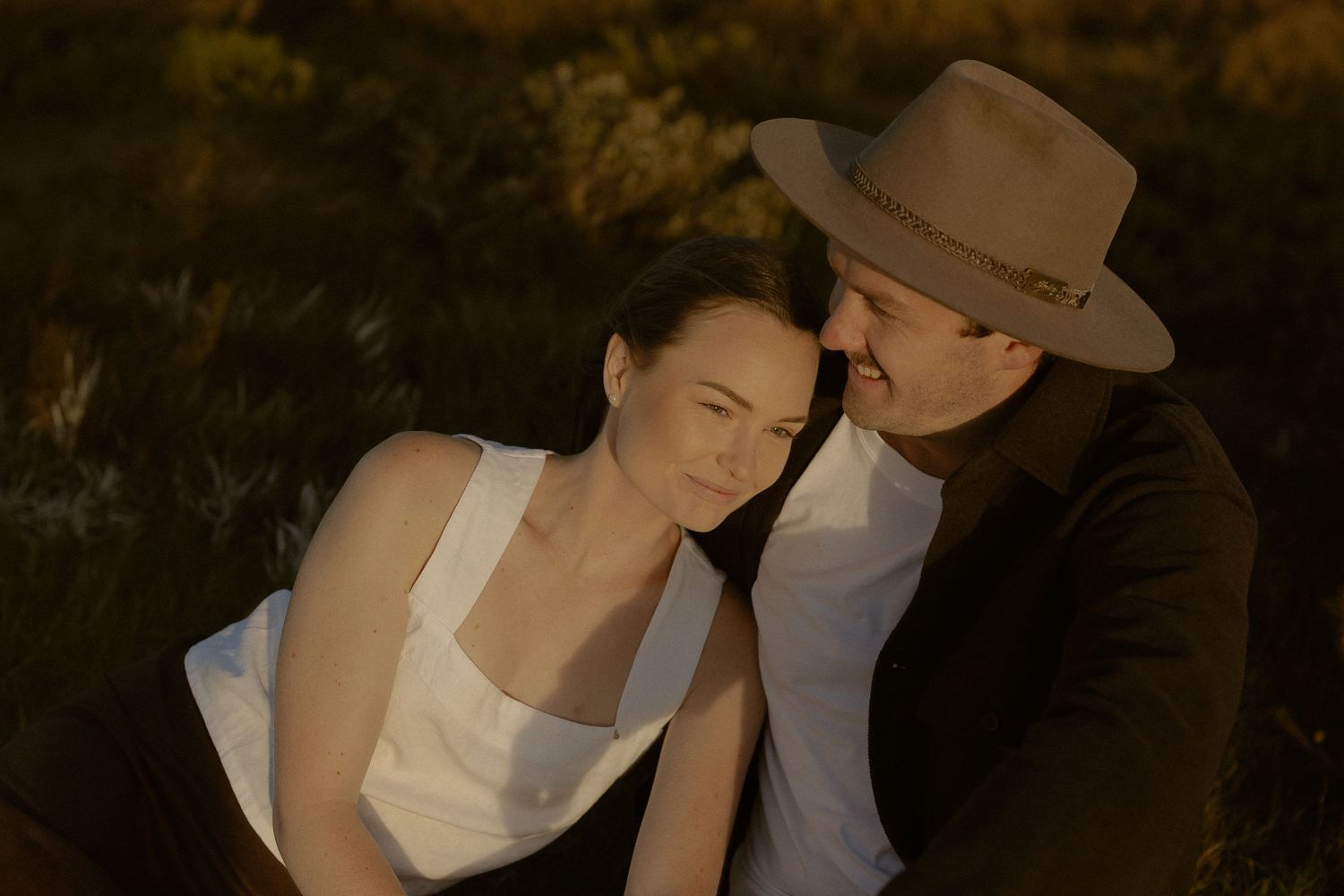A romantic couple in vintage-inspired clothing share an intimate moment at sunset in a dreamy outdoor setting.