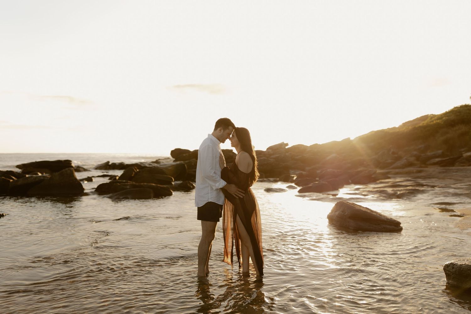 A couple embraces on a rocky beach at sunset with ocean waves lapping at their feet.