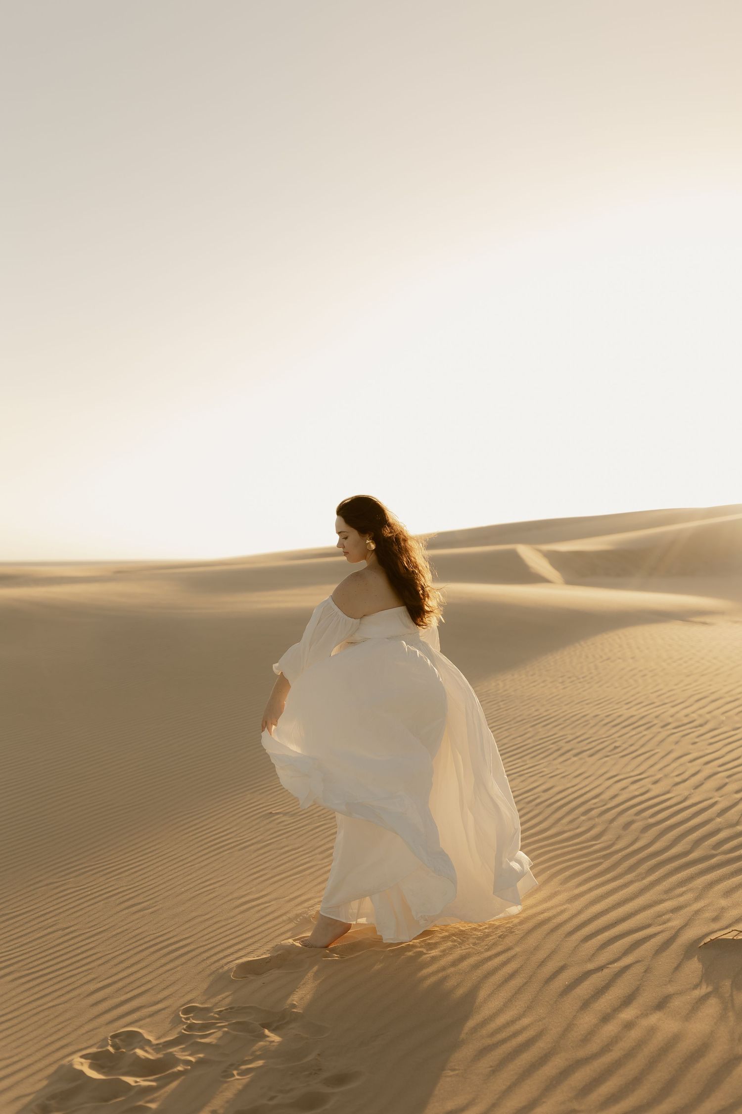A person in a flowing white dress walks gracefully across golden sand dunes in warm desert sunlight.