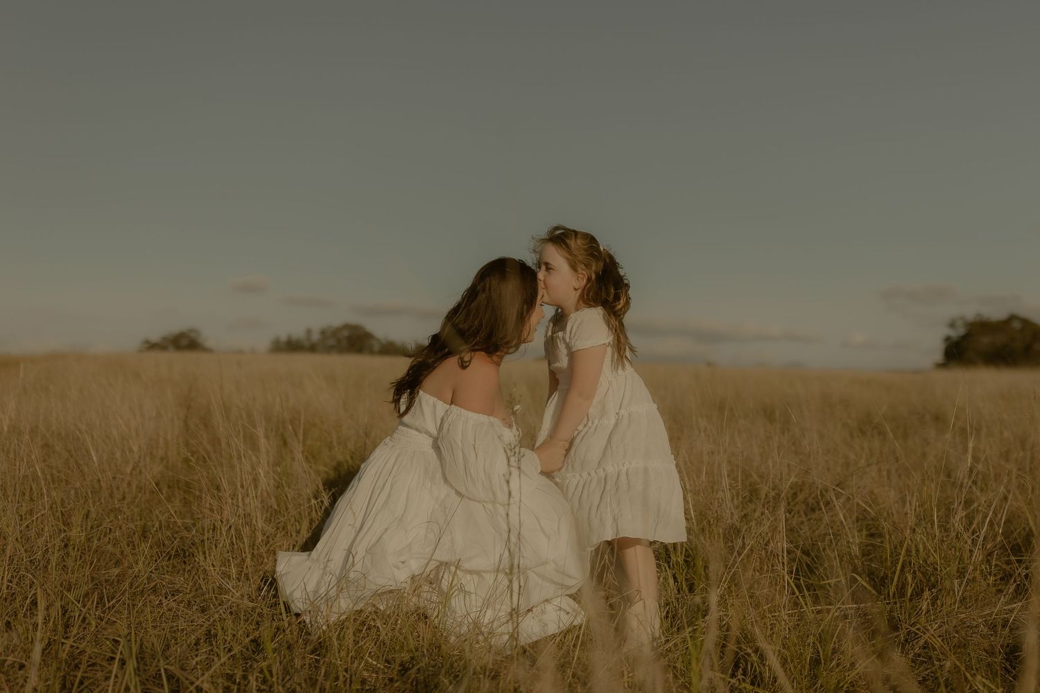 A couple shares an intimate moment in a golden wheat field at sunset while wearing light colored clothing.