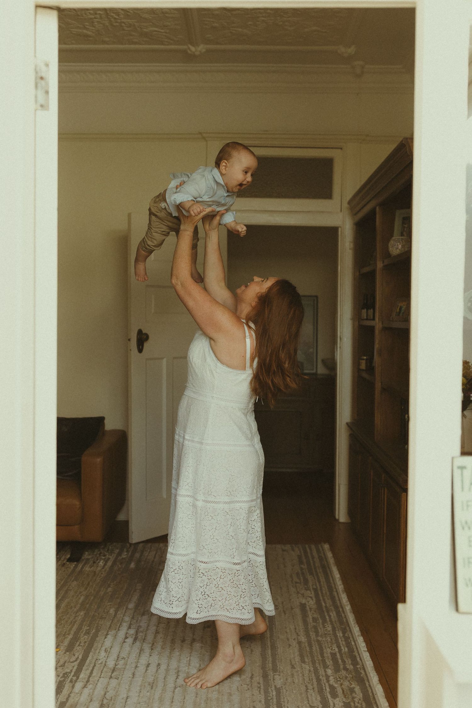 A tender moment captured in a hallway as a figure in a white dress lifts a small child playfully into the air.