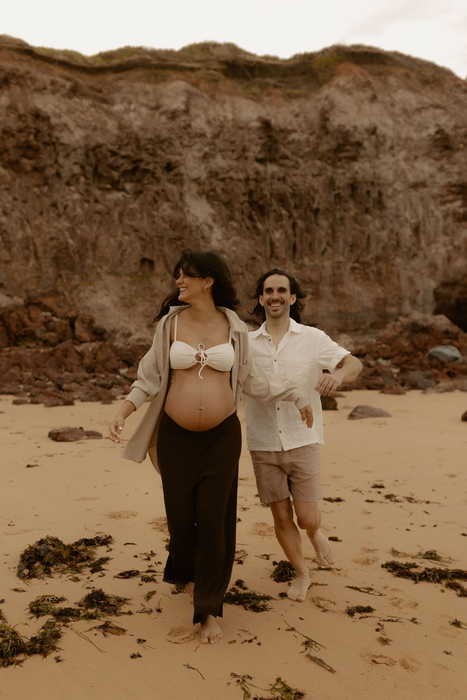 A sepia-toned beach photoshoot featuring two people walking along sandy shores near rocky cliffs.
