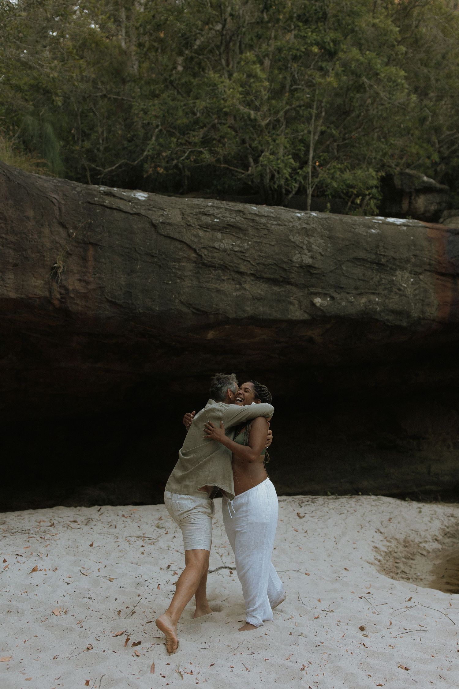 A couple poses playfully on white sand beneath a dramatic rock overhang in nature.