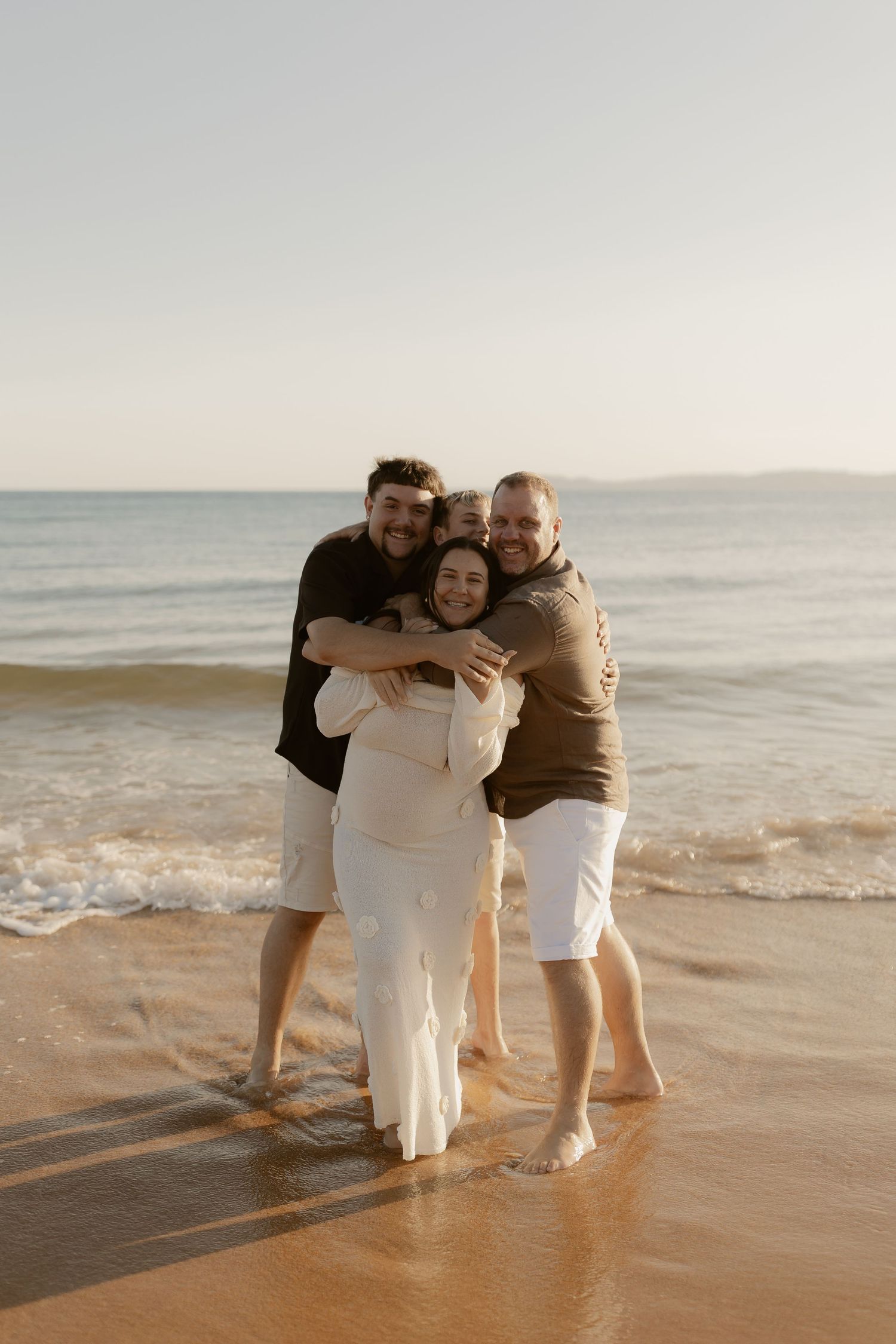 Three people embracing and laughing together on a sandy beach at sunset.