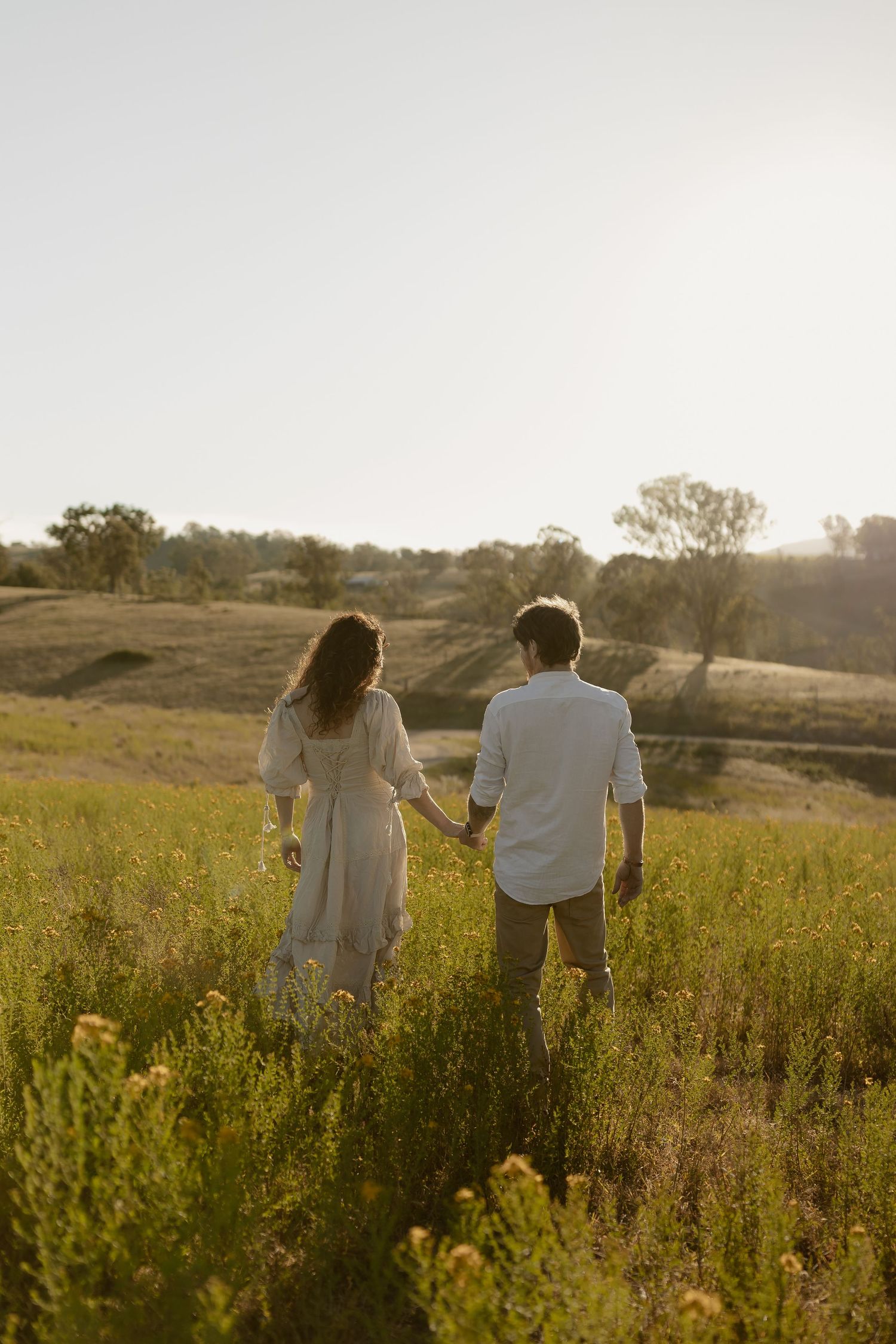A couple walks hand in hand through a sunlit meadow of tall grass and wildflowers at golden hour.