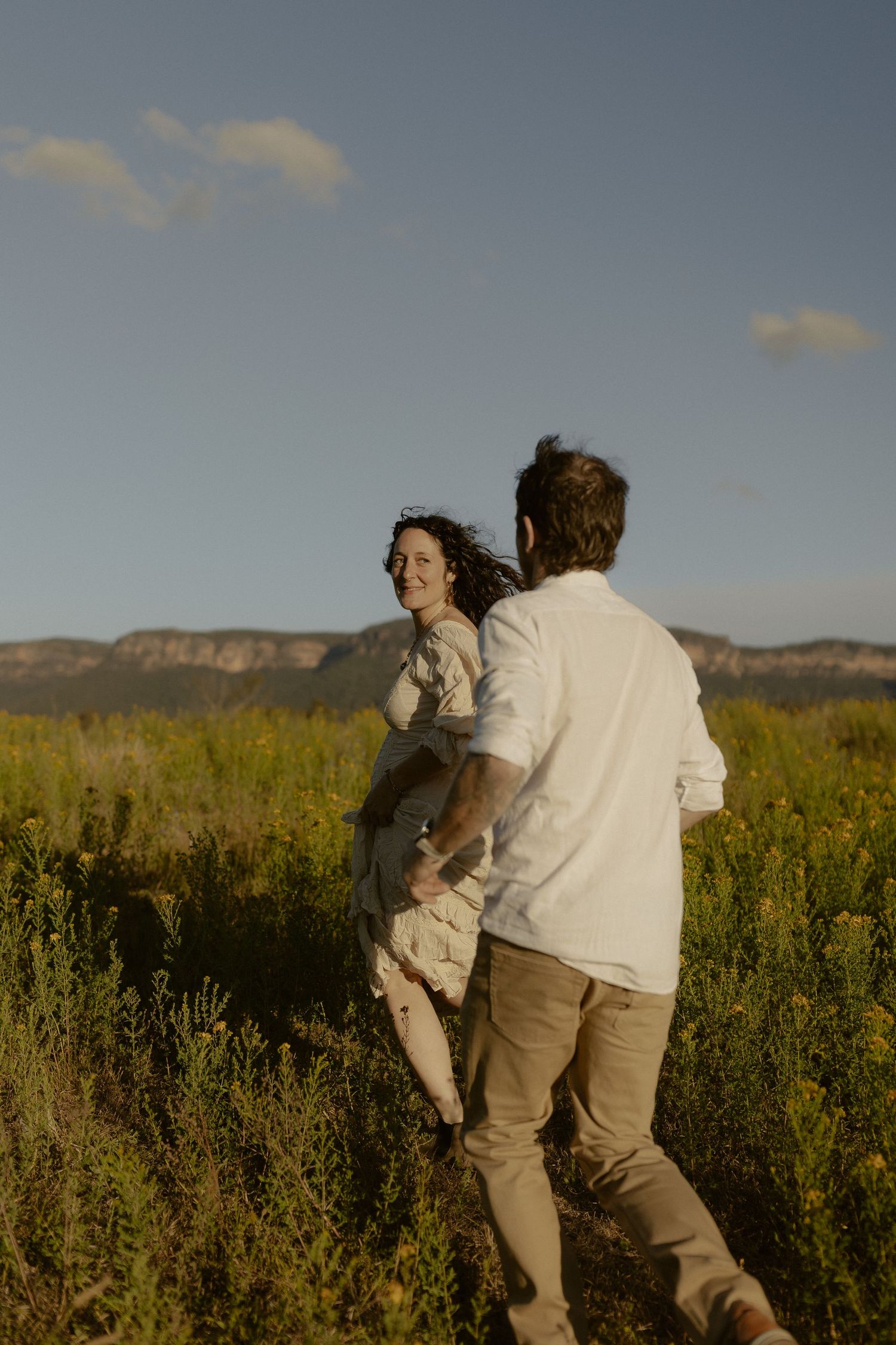Two people in light colored clothing embrace while standing in a field of yellow wildflowers at sunset.