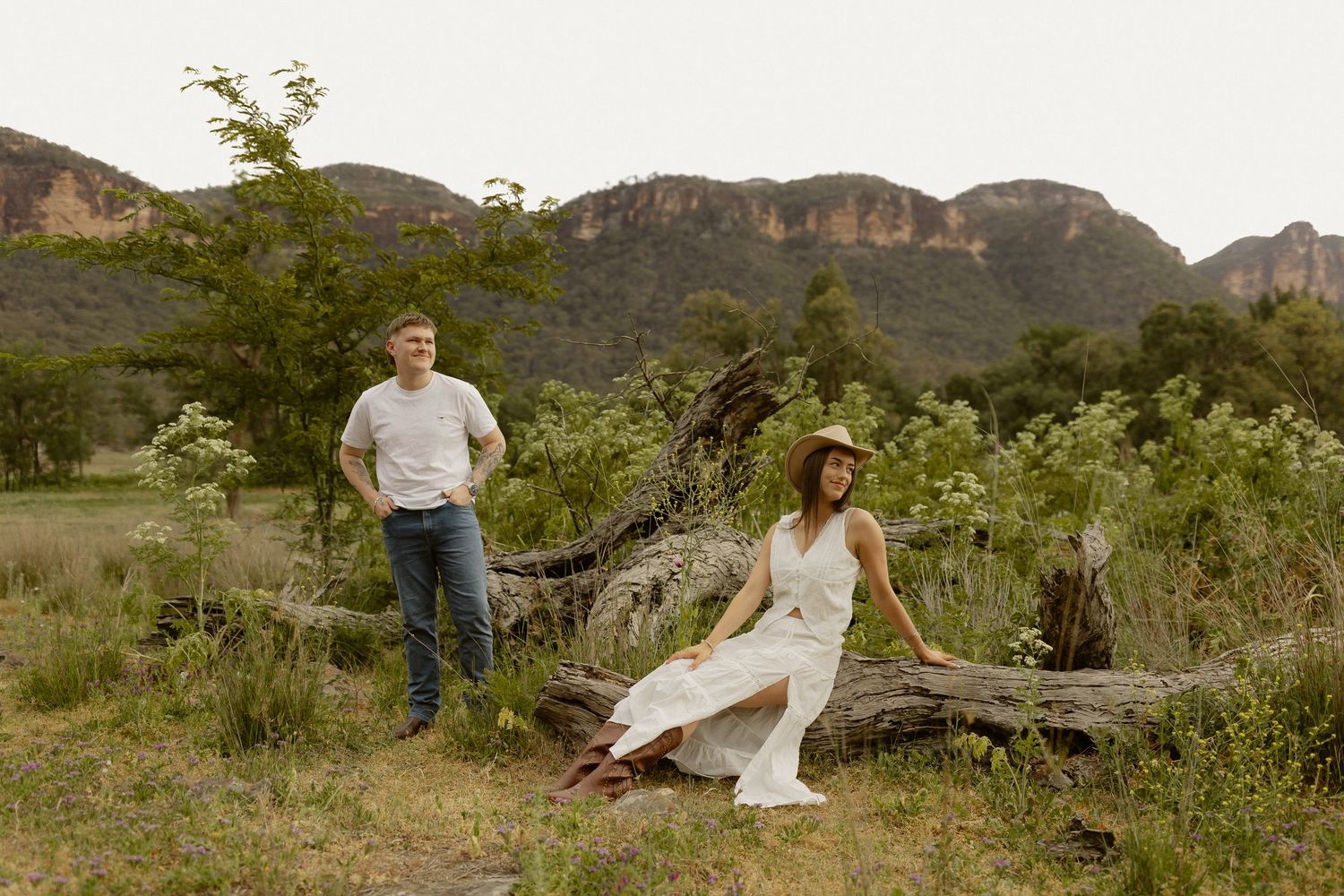 A couple poses in a scenic mountain landscape with rugged cliffs and green foliage in the background.