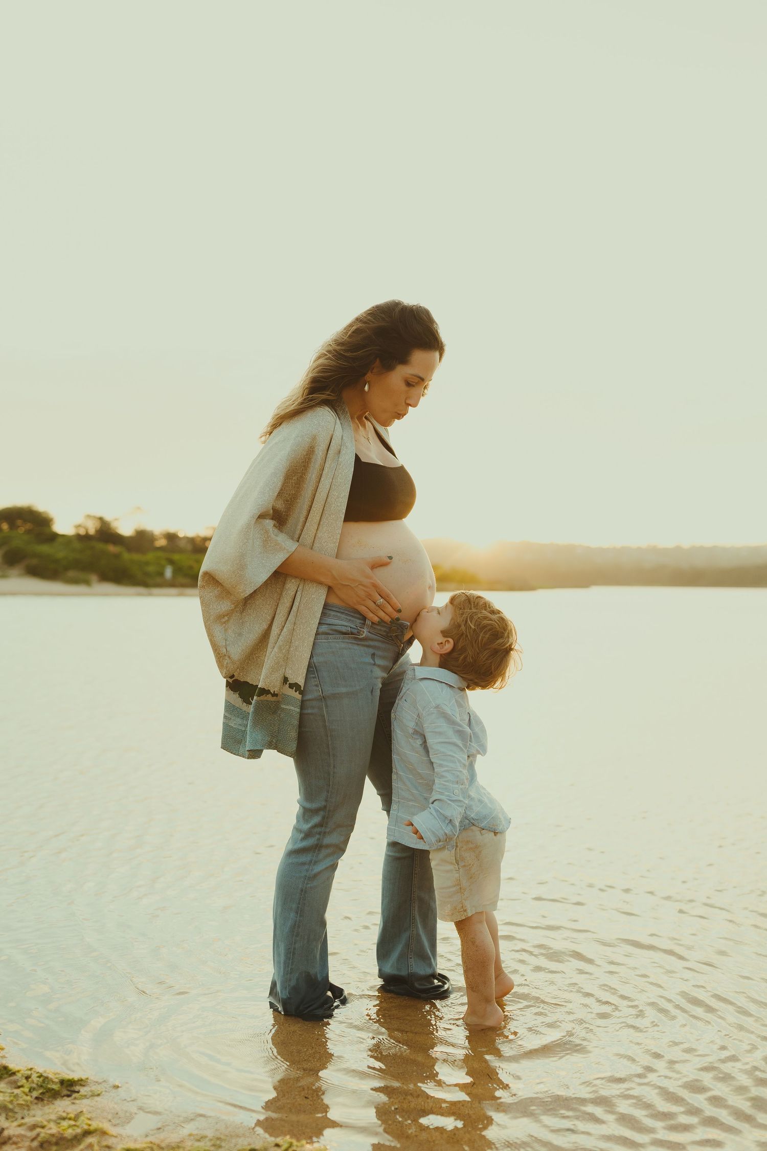 A parent and child wade together in shallow water at the beach during sunset.