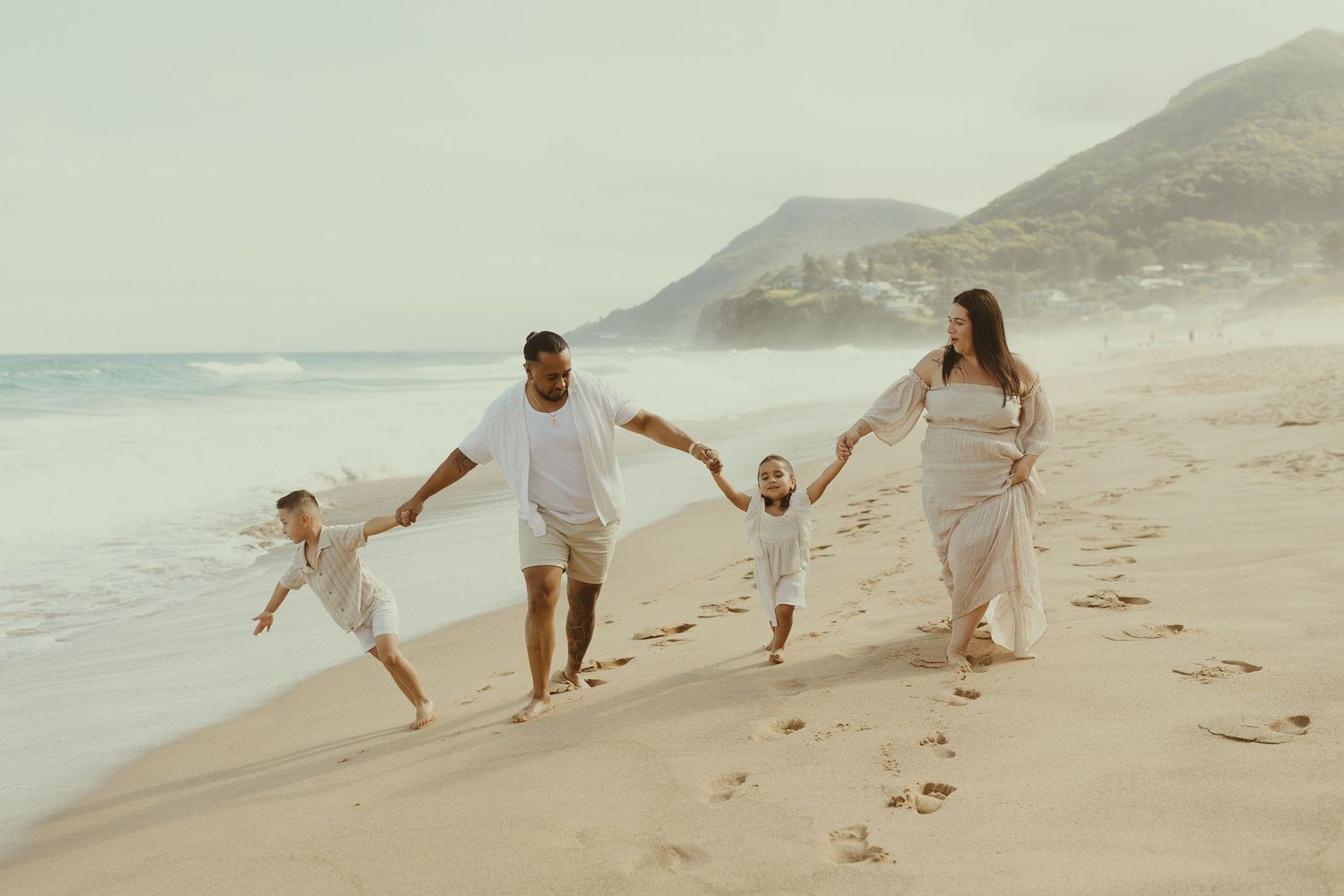 A family in white clothing holds hands while walking together on a sandy beach at sunset.