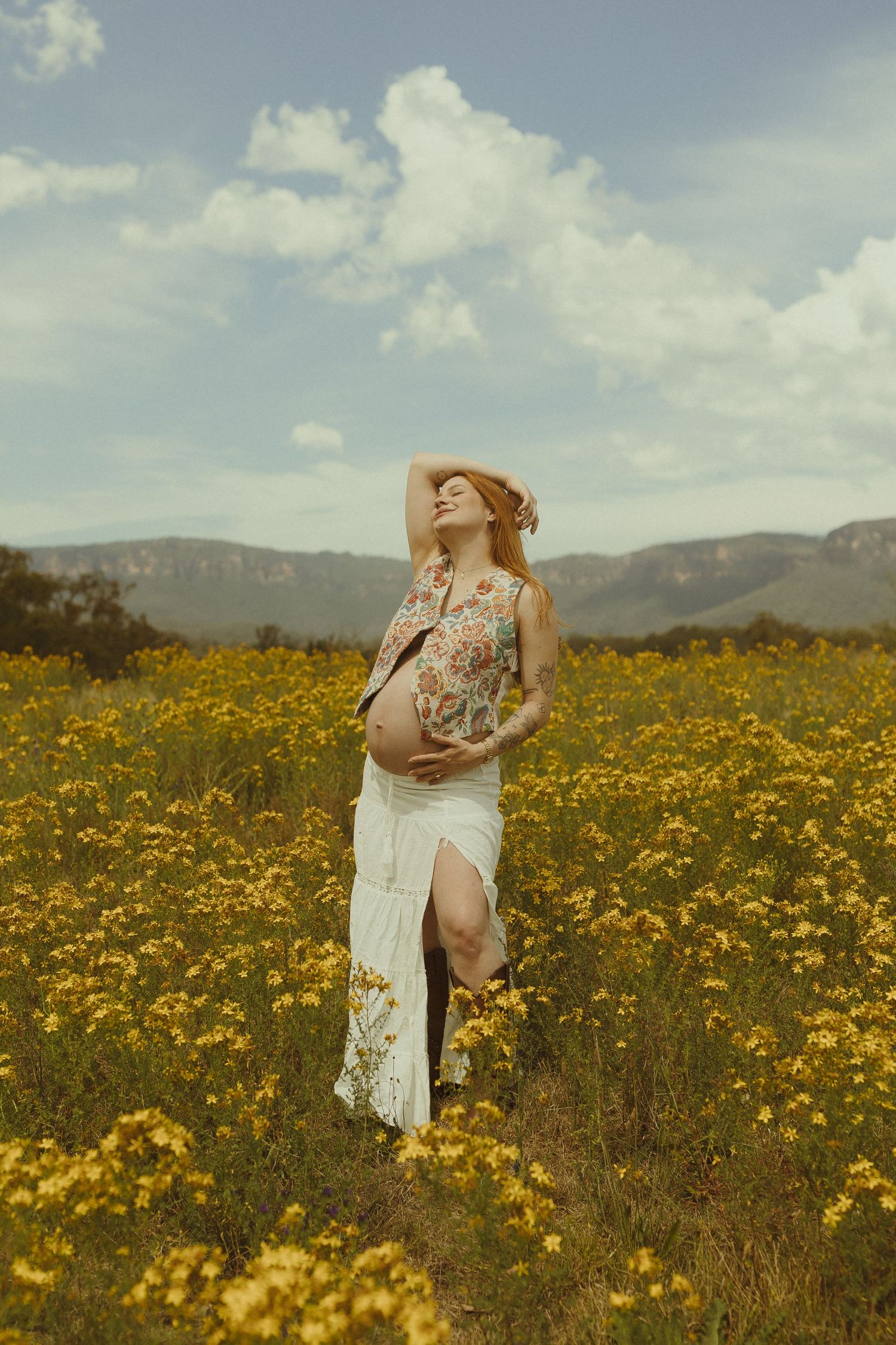 A figure in a white dress stands in a vibrant field of yellow wildflowers with mountains in the background.