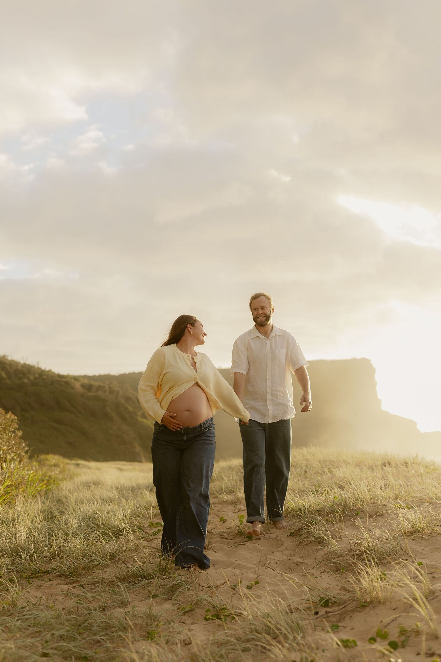 A couple walks together on grassy sand dunes at sunset in a romantic coastal setting.