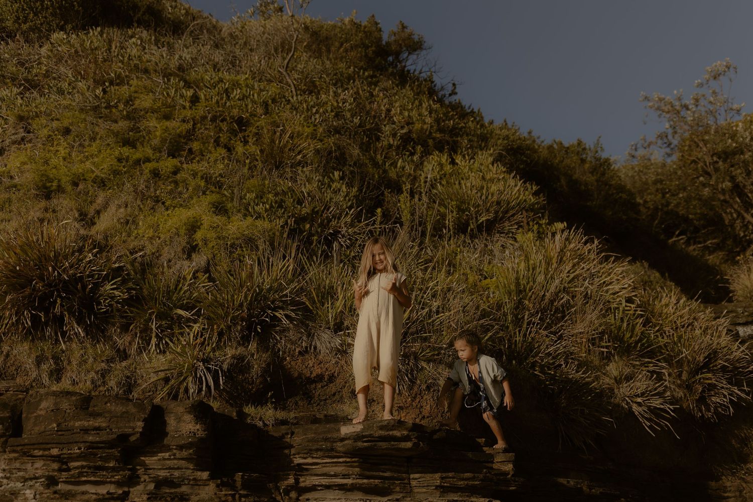 Two figures walk along a dirt path surrounded by golden sunlit brush and trees at sunset.