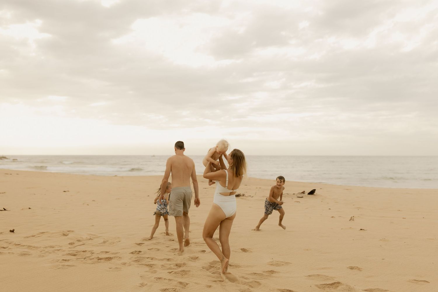 People and dogs playing together on a sandy beach during sunset with waves crashing in the background.