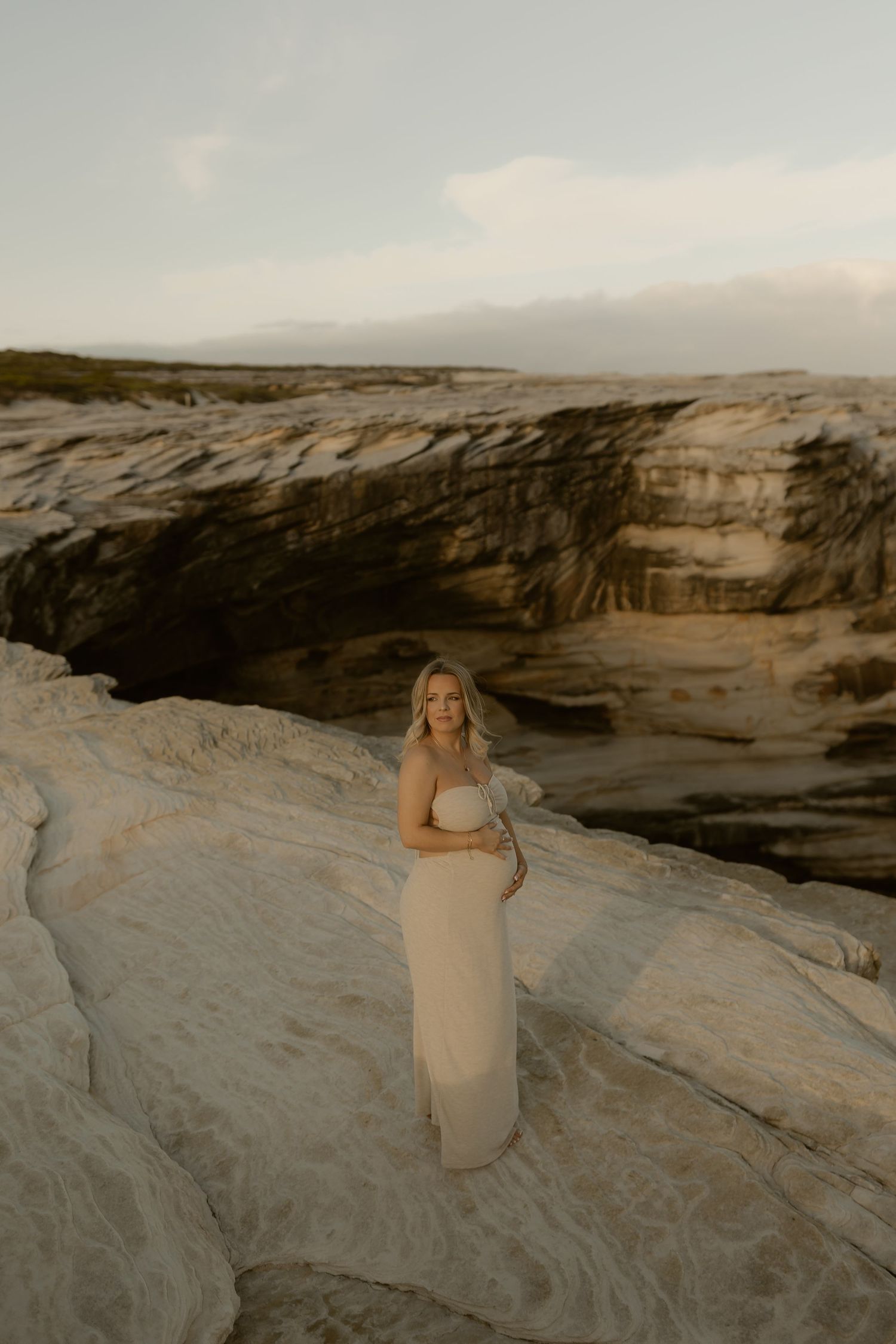 Person in flowing dress poses on dramatic rocky cliffs at sunset with ethereal lighting.
