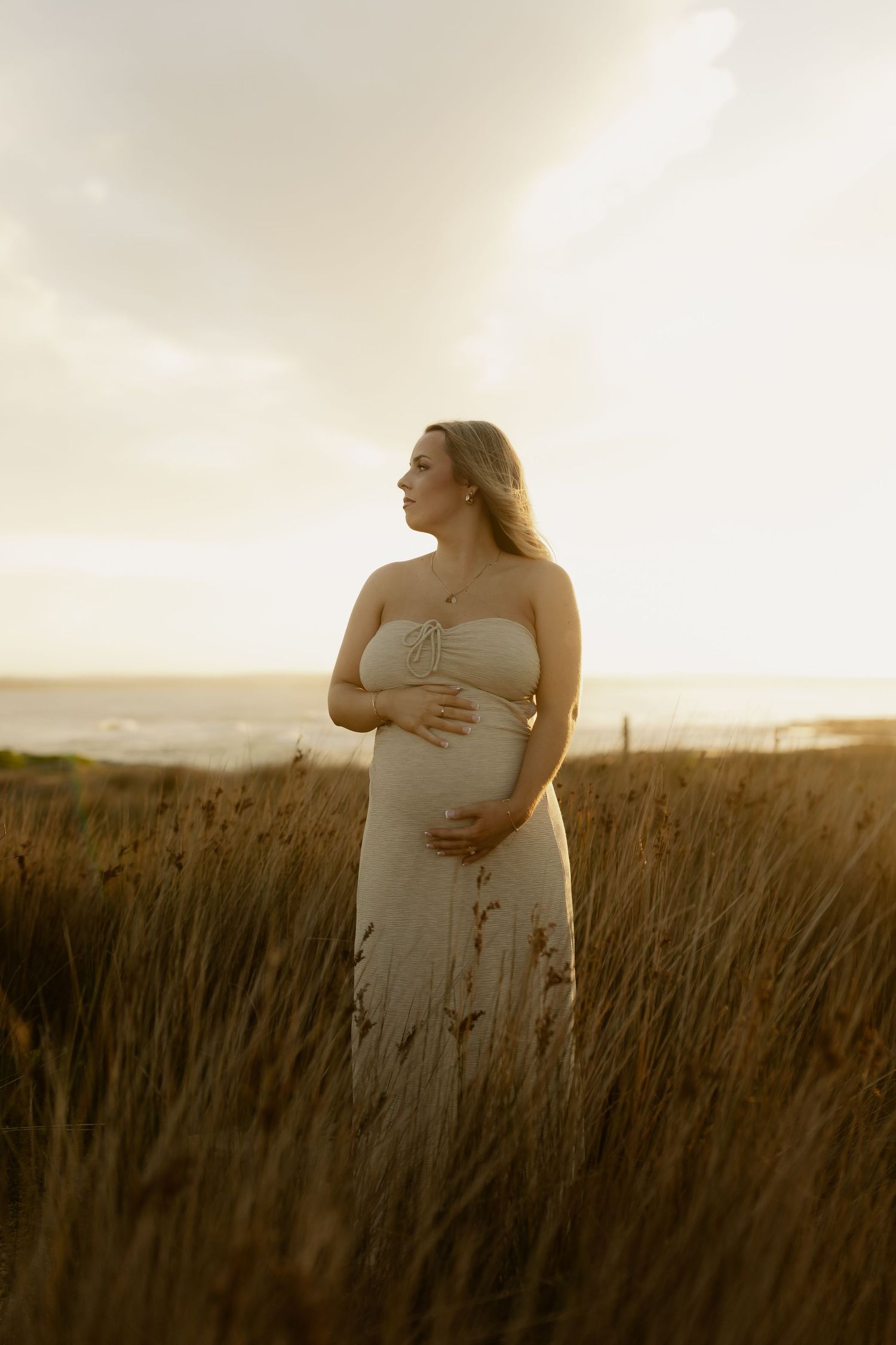 A person in a white dress stands in a field of tall grass during sunset, cradling their belly.