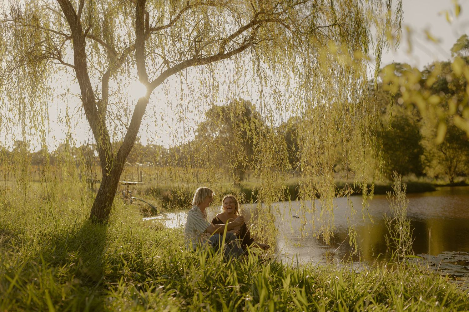 Dreamy vintage scene of figures by a tranquil pond with drooping willow trees in warm afternoon light.