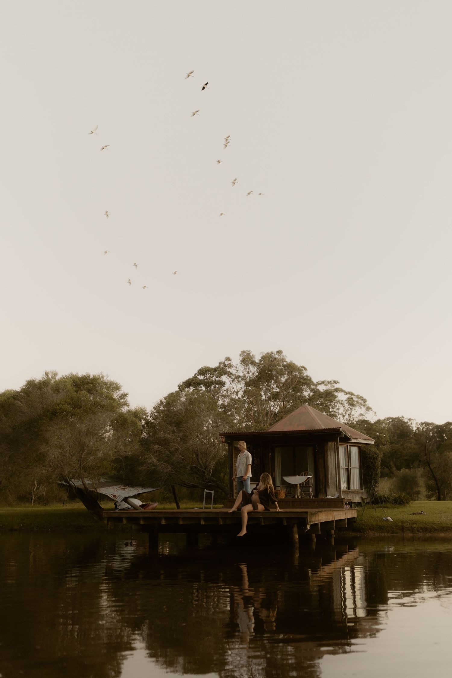 Rustic wooden dock extends over calm water at sunset with small boathouse structure visible in the distance.