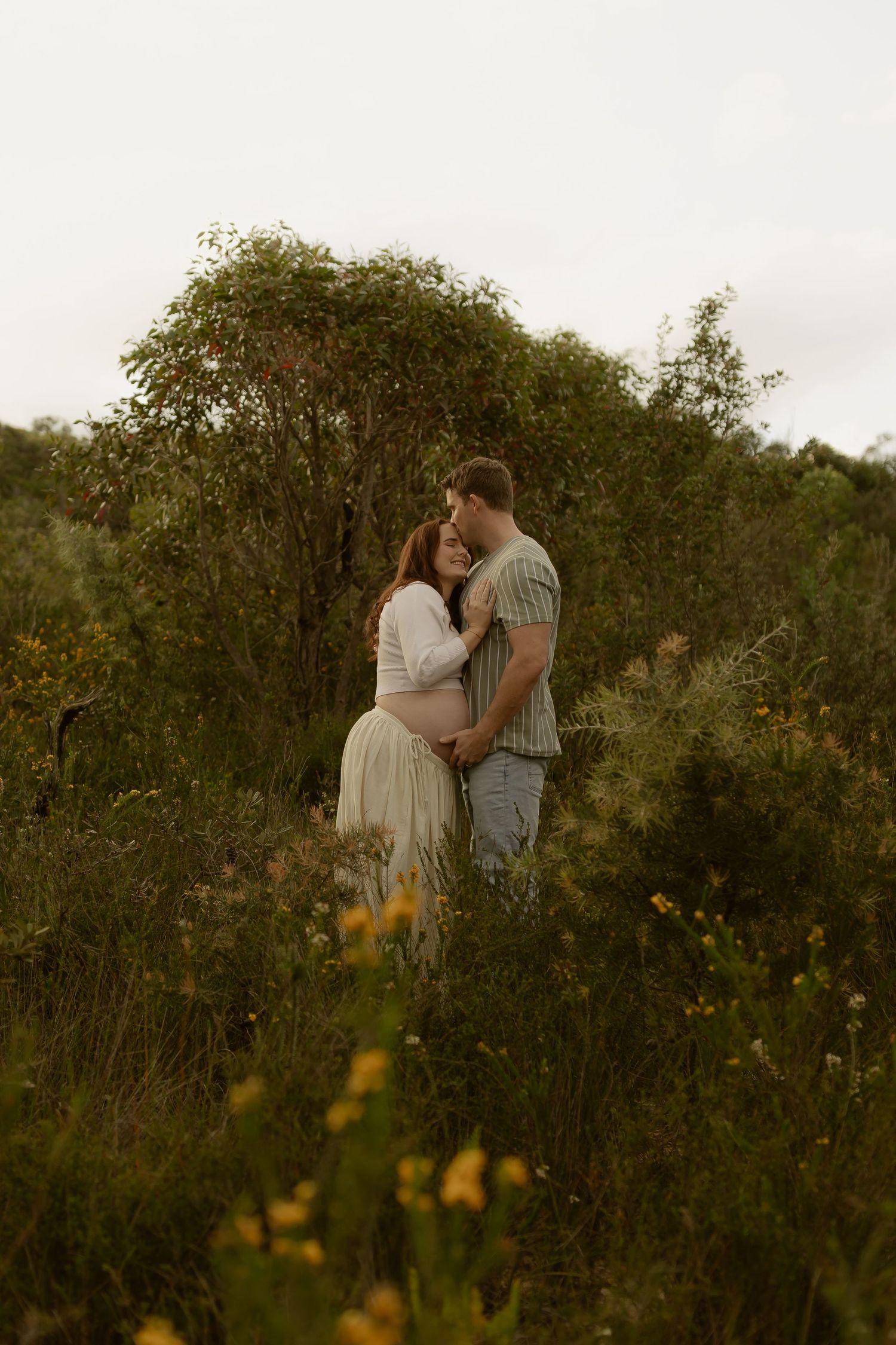 Couple embracing in a lush green field, maternity photo shoot with wildflowers and trees in natural outdoor setting.