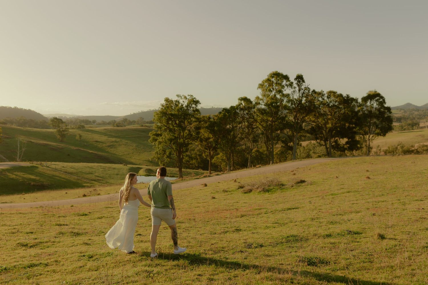 A bride and groom walk hand in hand across a lush green hillside with scenic rolling countryside views at golden hour.
