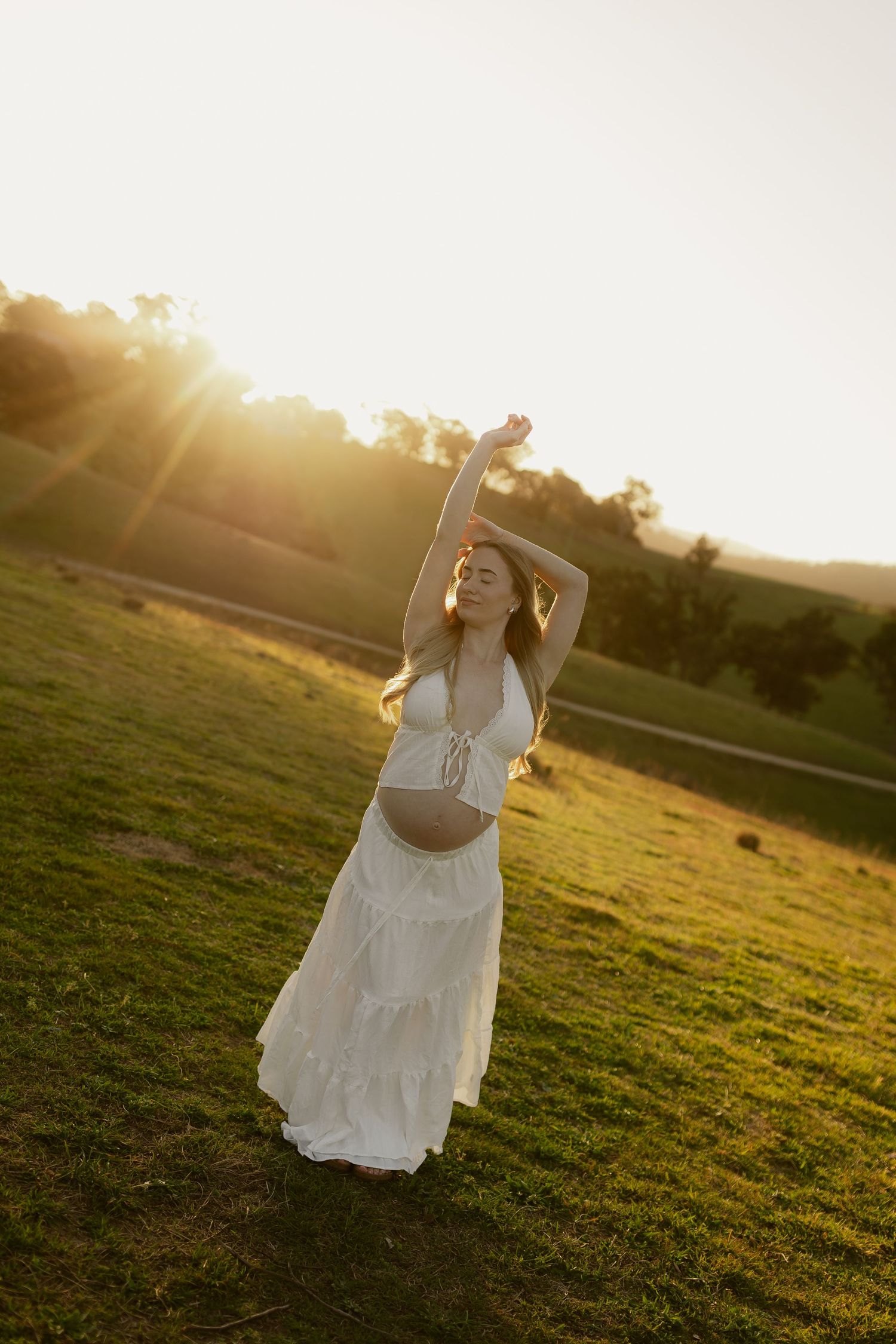 Pregnant woman in white crop top and maxi skirt posing at golden hour in a green field.