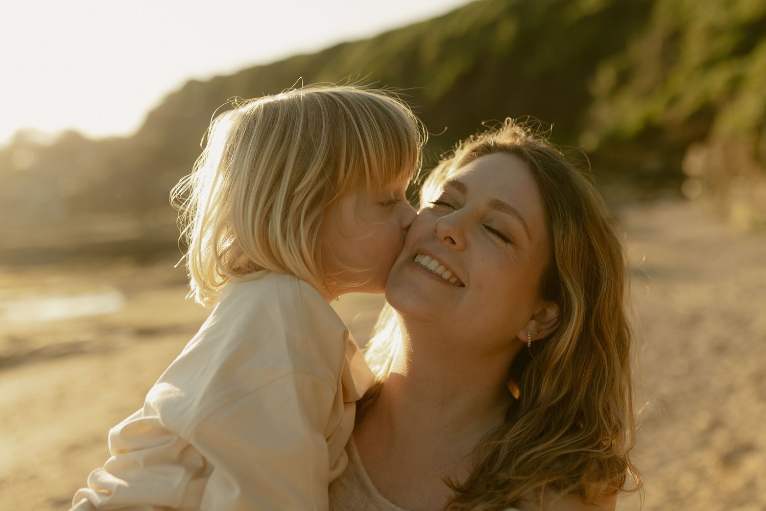 A young child kisses a smiling woman on the cheek at a sunny beach during golden hour.