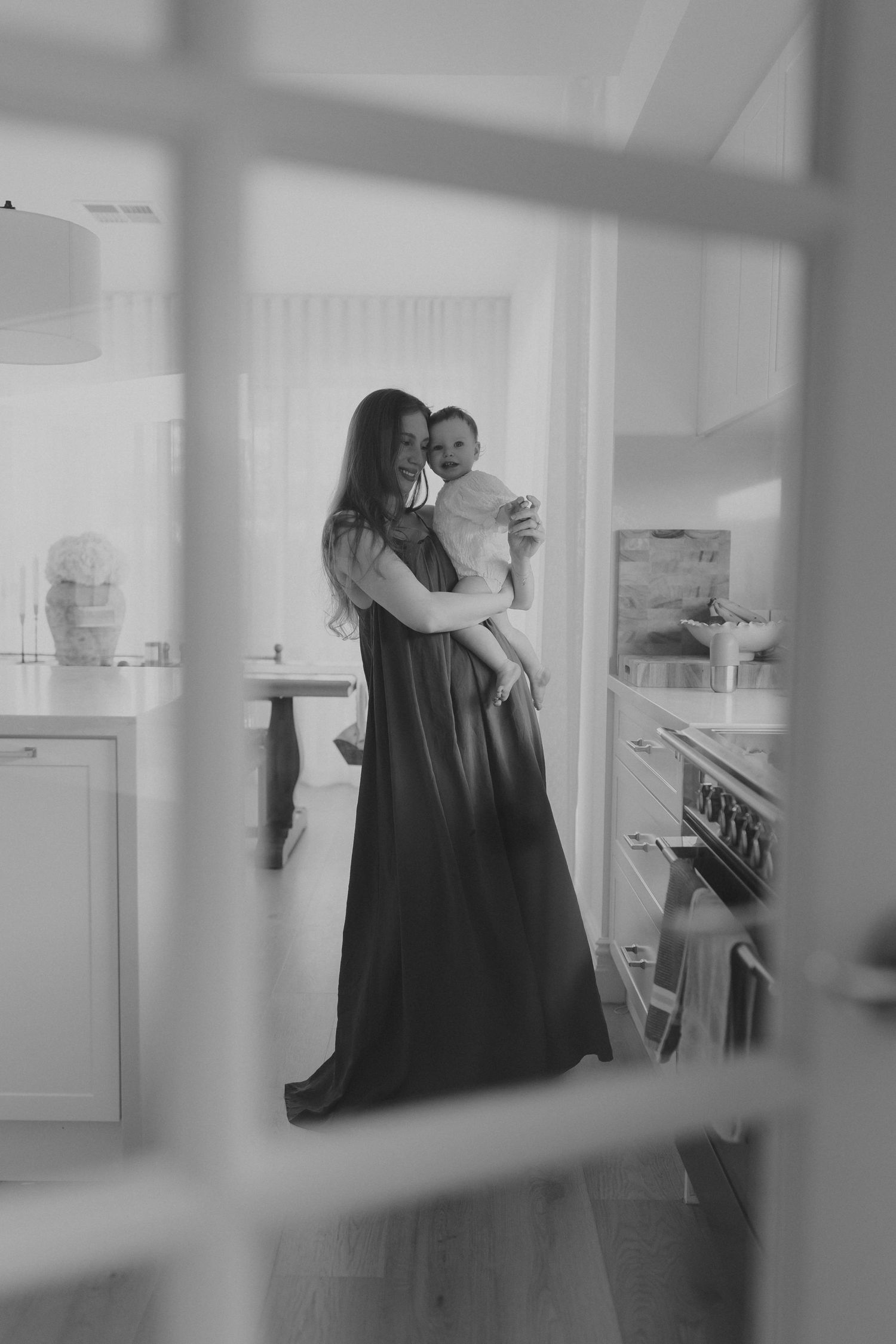 Black and white photo of a mother in a long dress holding her baby in a bright modern kitchen.