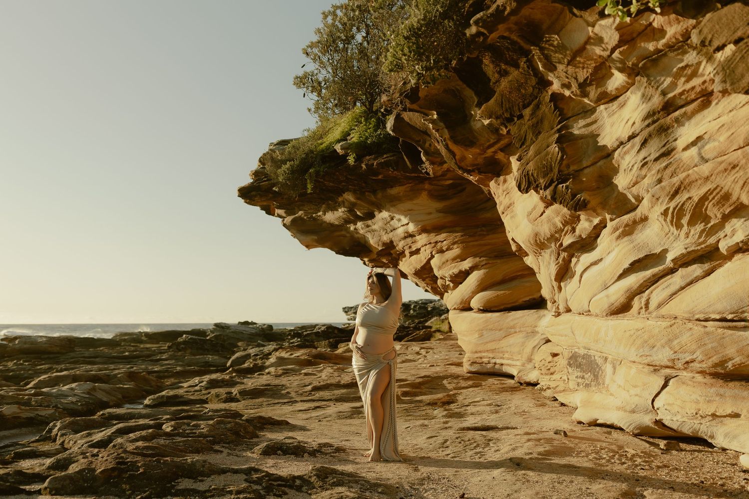 Woman standing beside stunning sandstone cliffs on a rocky beach at golden hour.