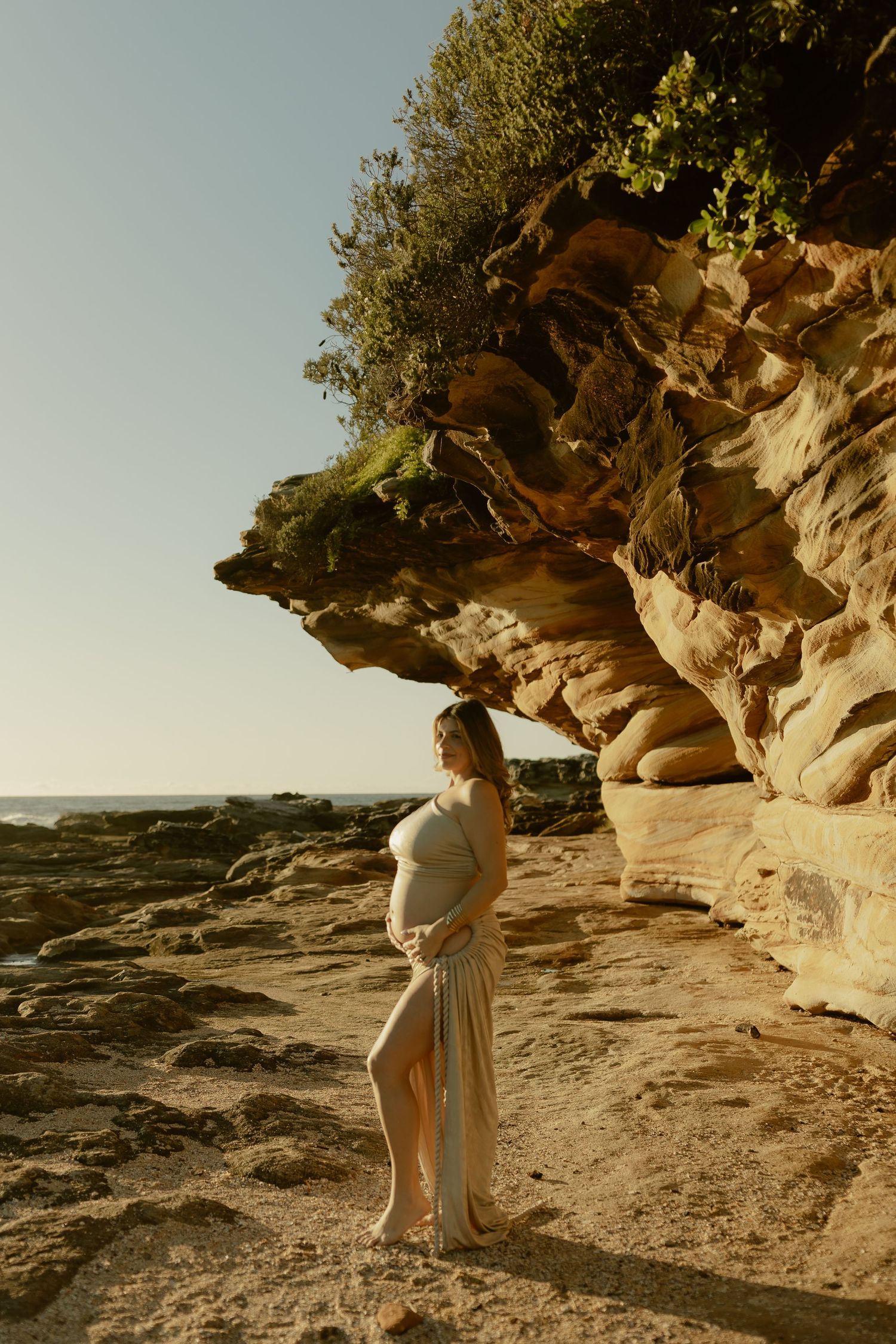 Pregnant woman in flowing white dress cradling baby bump beside golden sandstone cliffs at beach during sunset.