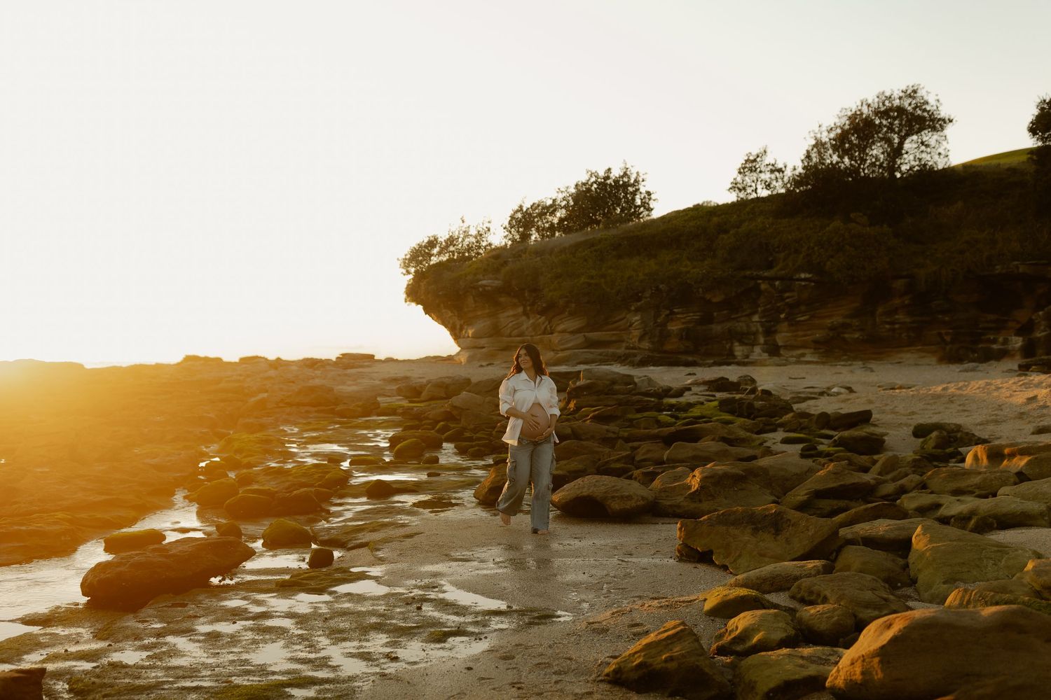 Woman standing among rocks on a scenic beach at golden hour with a glowing sunset behind coastal cliffs.