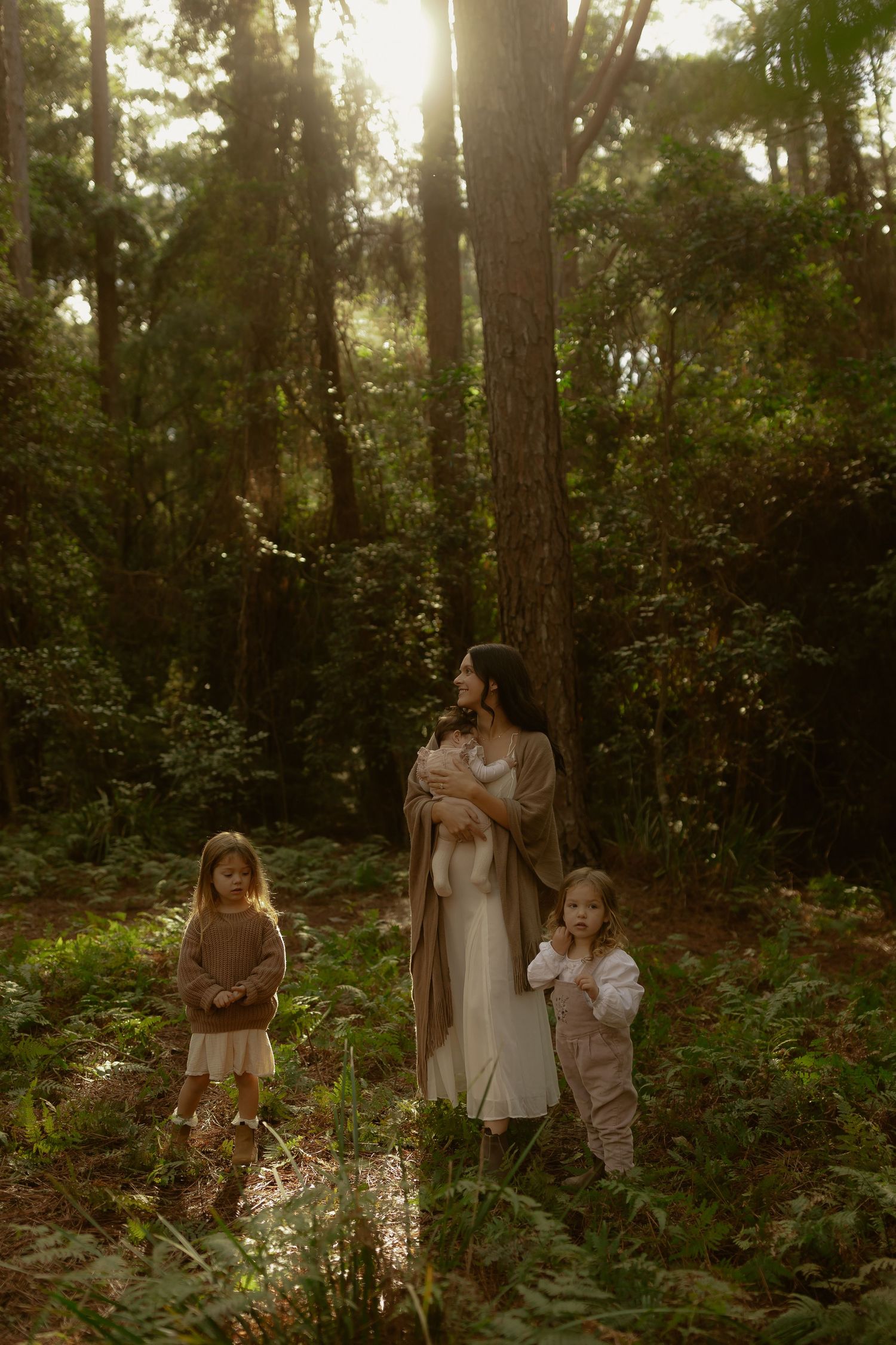 Mother holding newborn with two children in golden sunlit forest clearing.