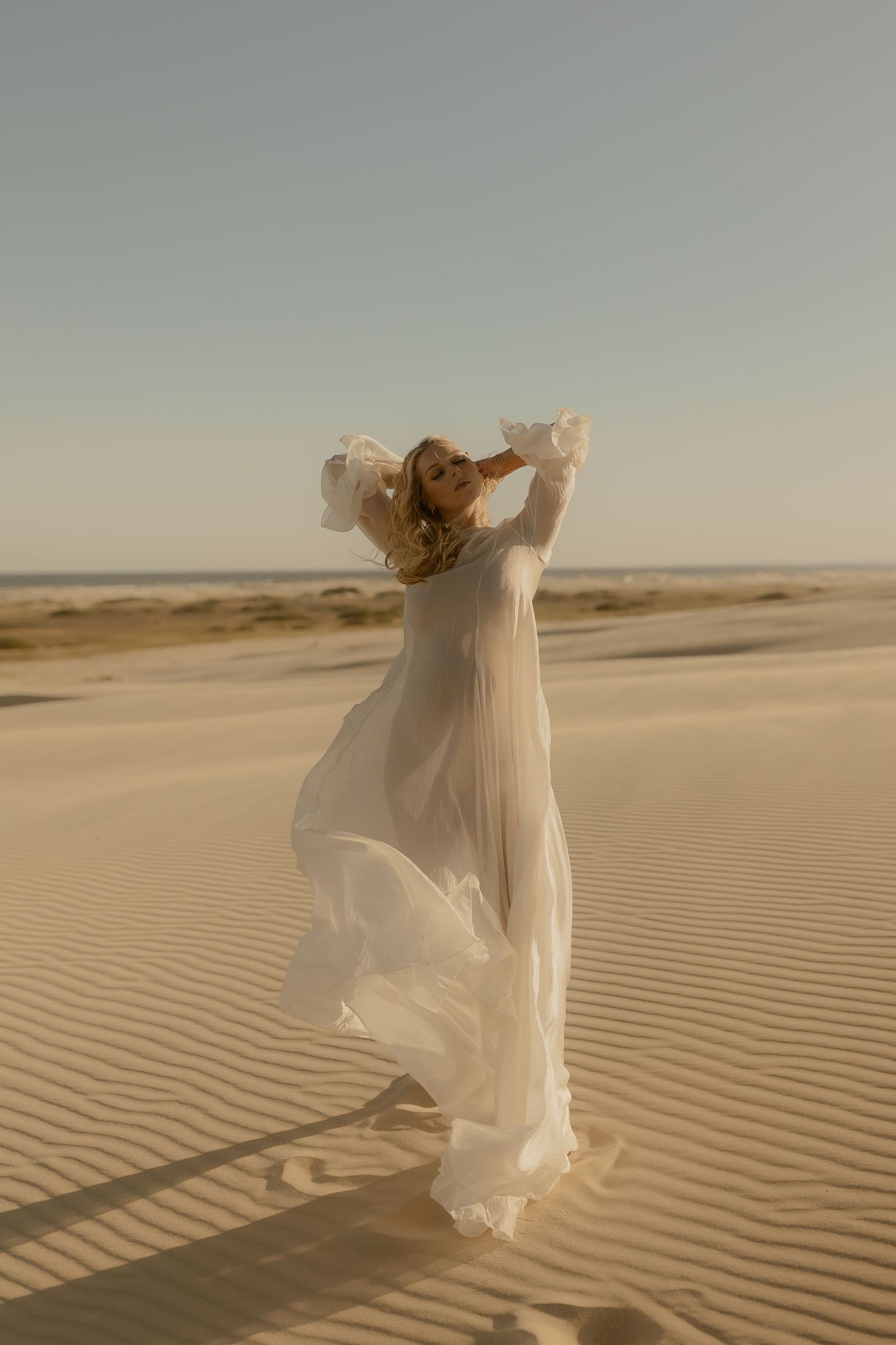 Woman in flowing white dress standing on desert sand dunes with arms raised against clear sky.