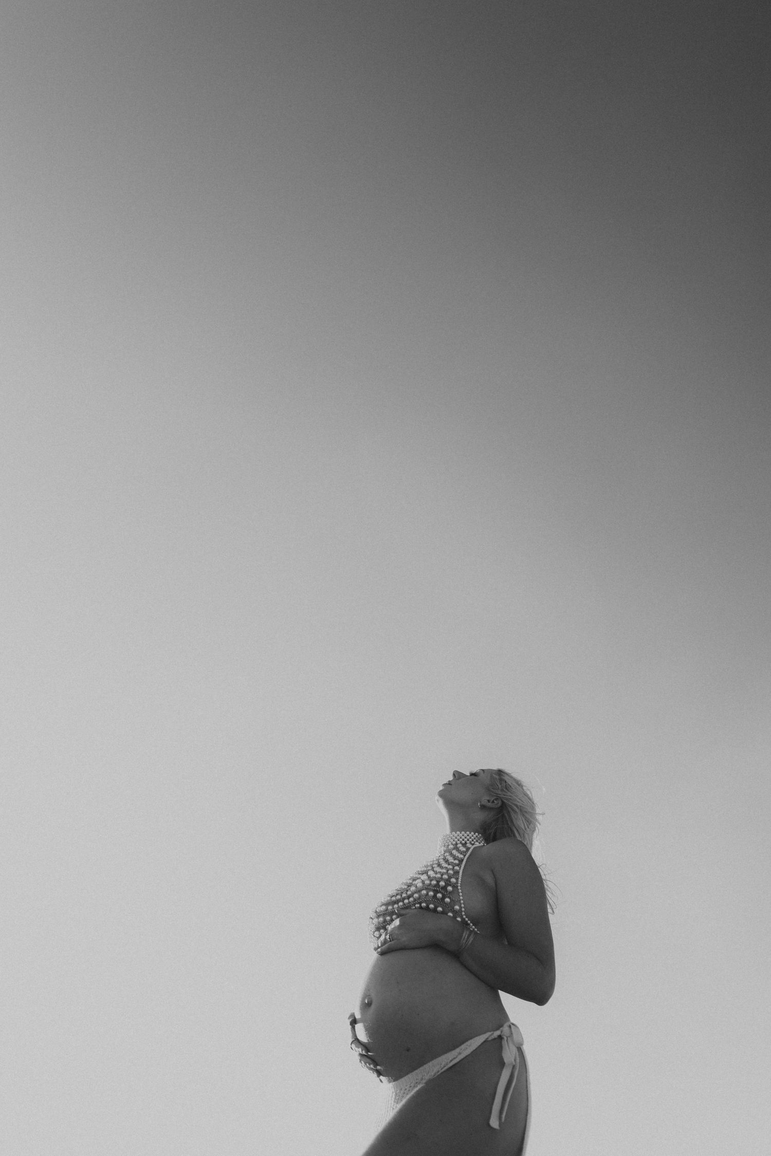 Black and white photo of pregnant woman in bikini looking up at sky outdoors.