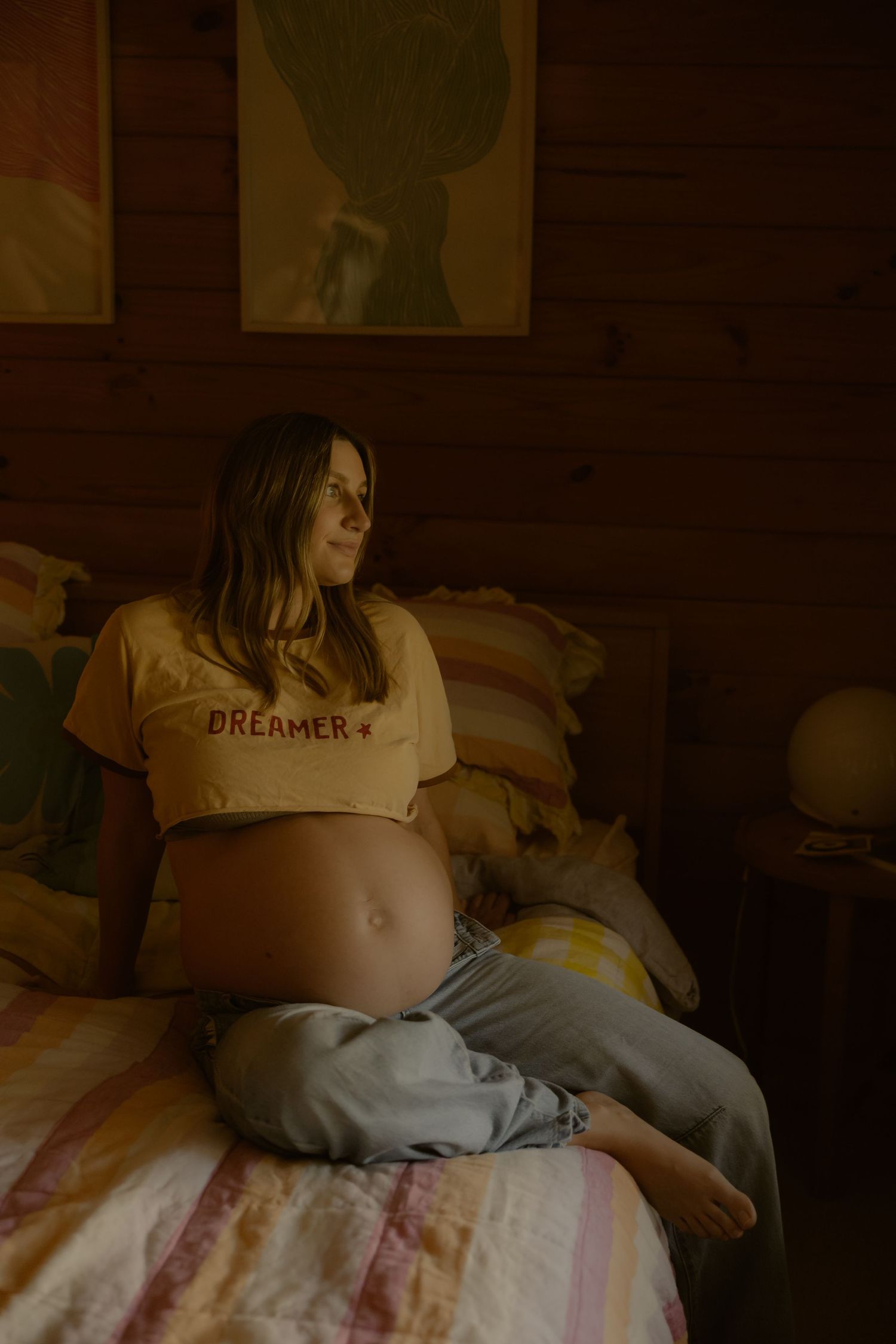 Pregnant woman in yellow Dreamer crop top sitting cross-legged on bed in warmly lit wooden cabin bedroom.