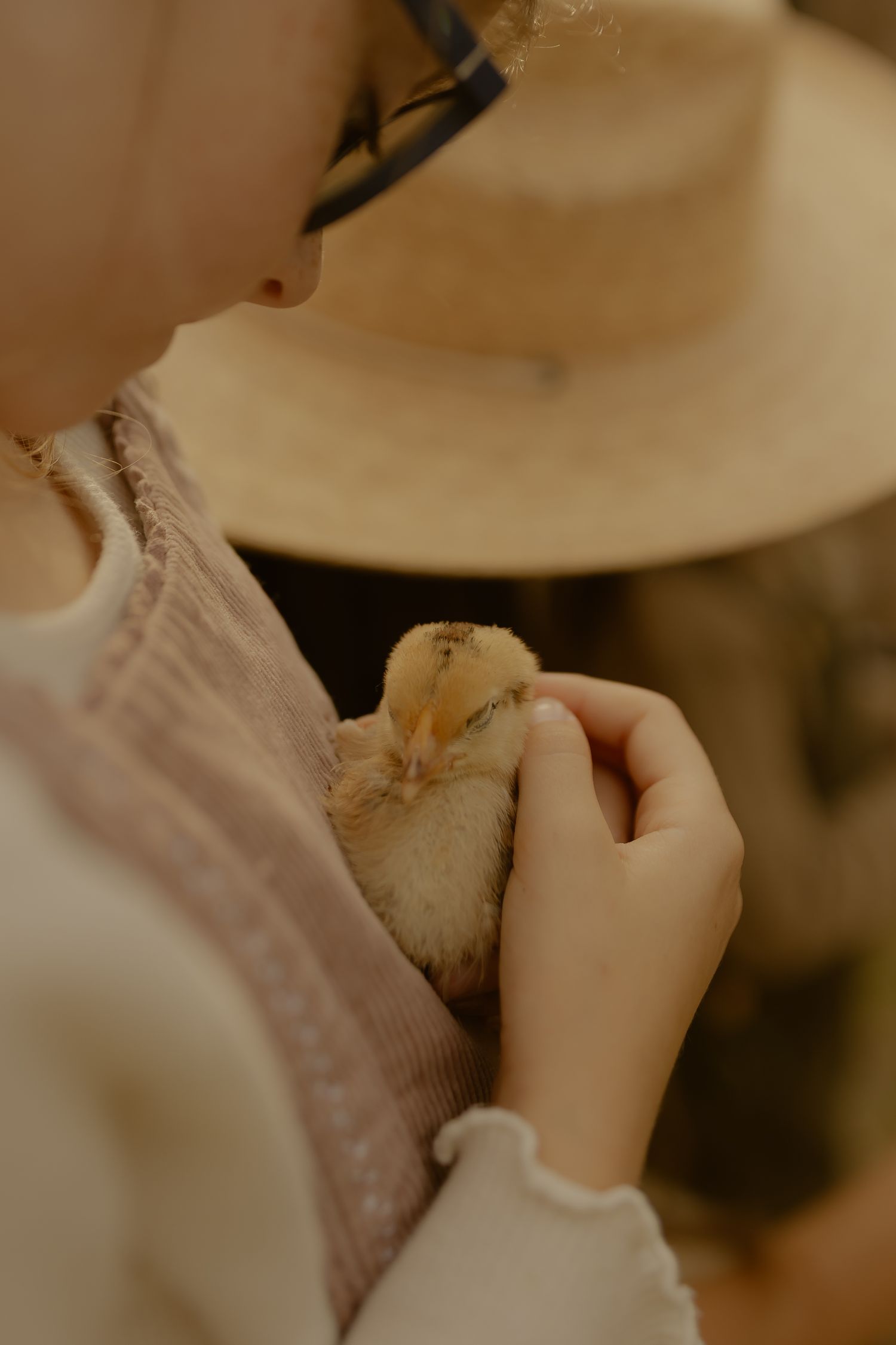 A young child in glasses gently holds a tiny yellow baby chick close to their chest.