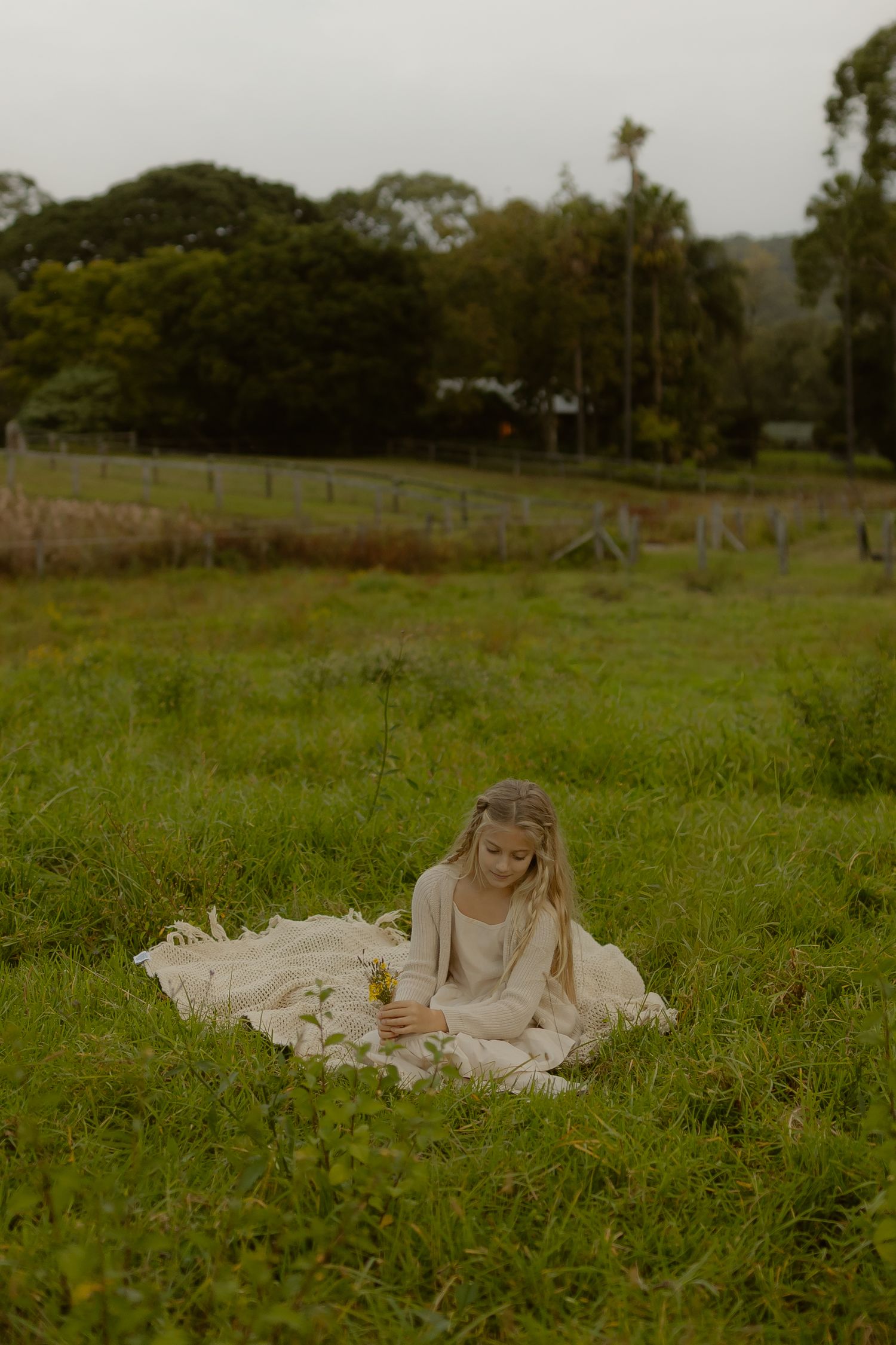 Young girl in cream dress sitting on blanket in lush green field holding a flower.