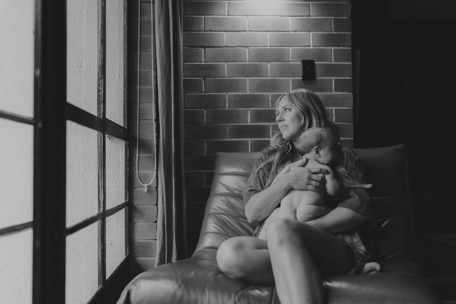 Black and white photo of a woman sitting in a leather chair holding a baby, gazing out a window beside a brick wall.