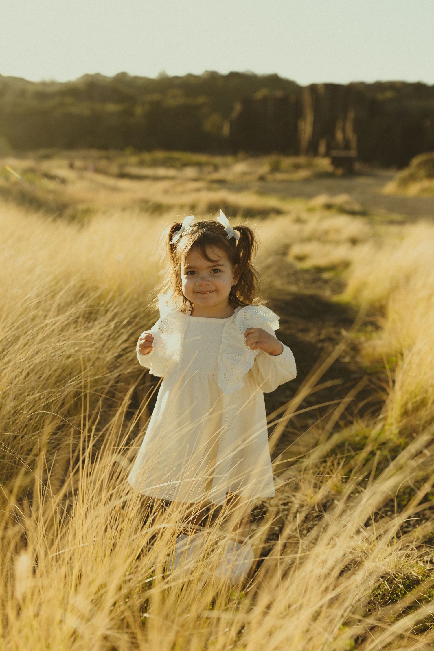 White dress-clad figure stands in golden grass field at sunset with mountains in background.