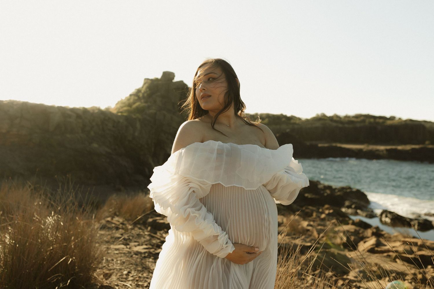 A maternity photo shows a woman in a white ruffled dress posing against a sunset waterfront backdrop.
