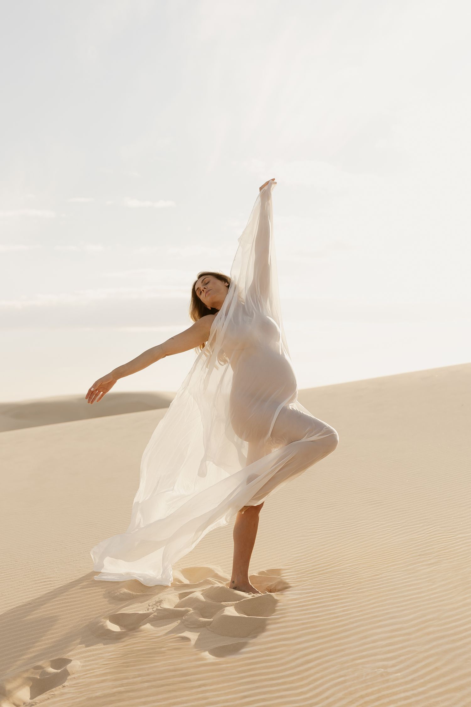 Figure in flowing white fabric dances gracefully across desert sand dunes in ethereal desert photoshoot.