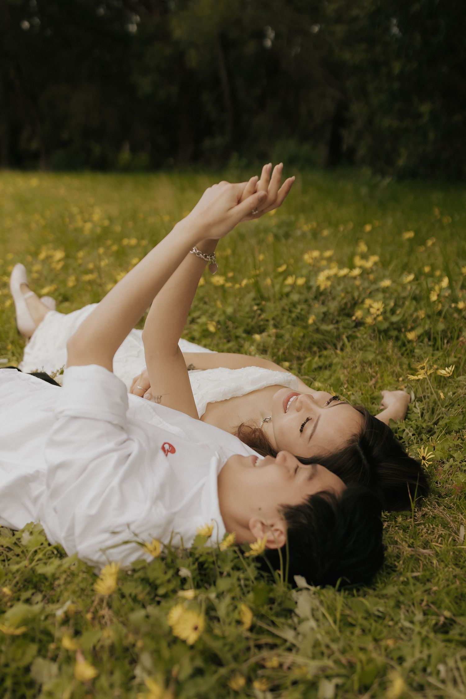A couple relaxes on grass reaching their hands up toward the sky during an outdoor photoshoot.