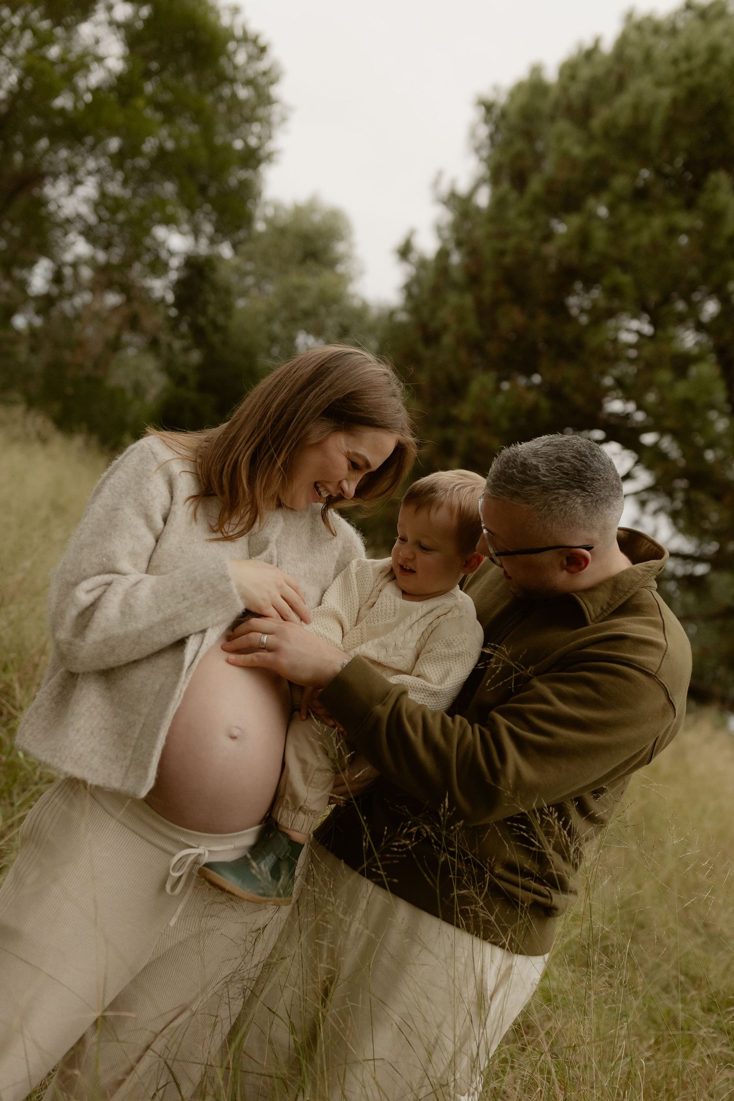 Expecting couple sharing a tender moment with their toddler in a lush green field during maternity shoot.