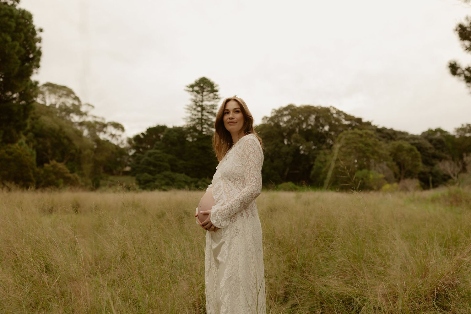 Pregnant woman in white lace dress standing in tall grass field with trees in background.