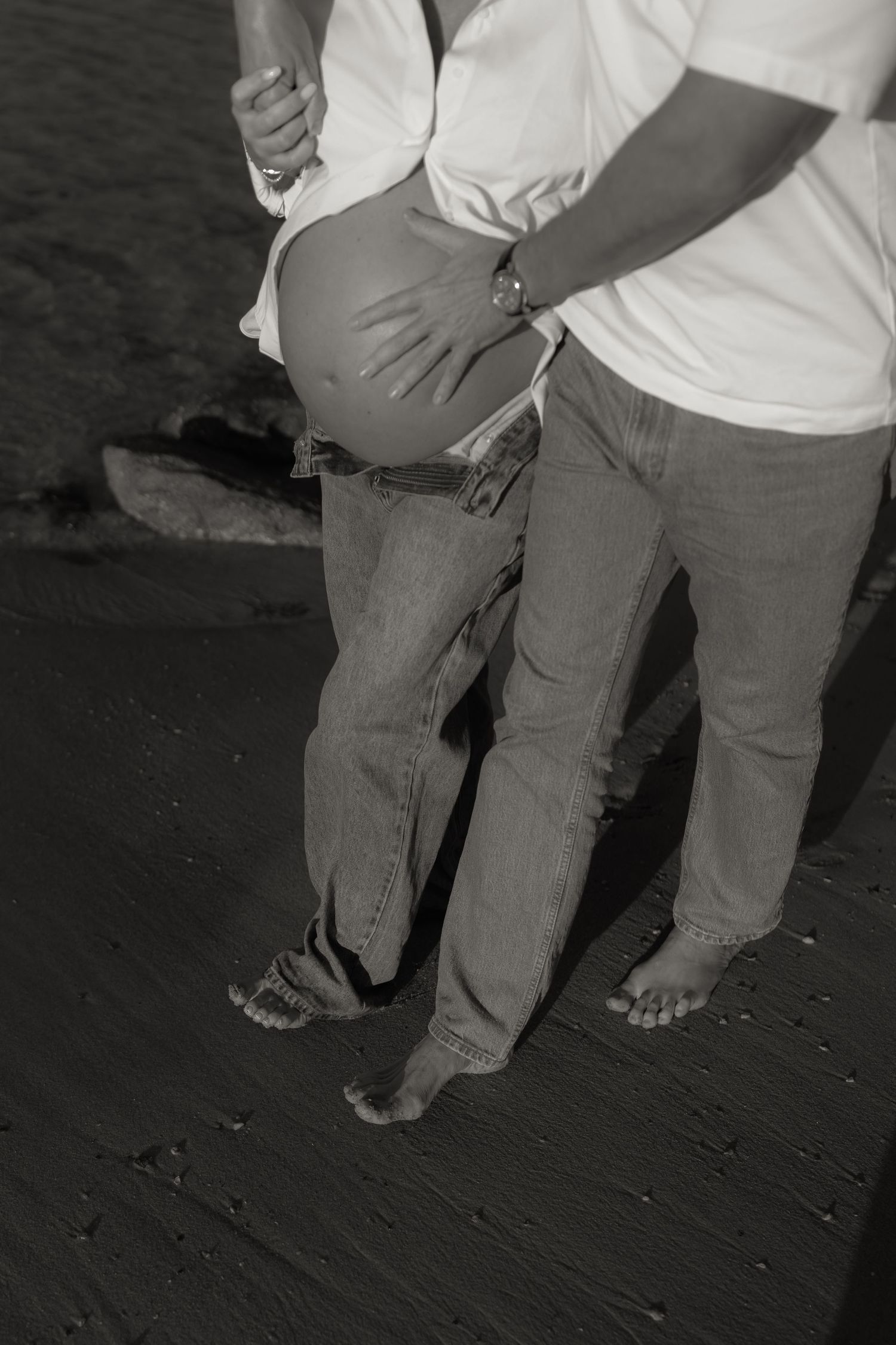 Black and white close-up of a couple barefoot on a beach, man gently holding pregnant belly.
